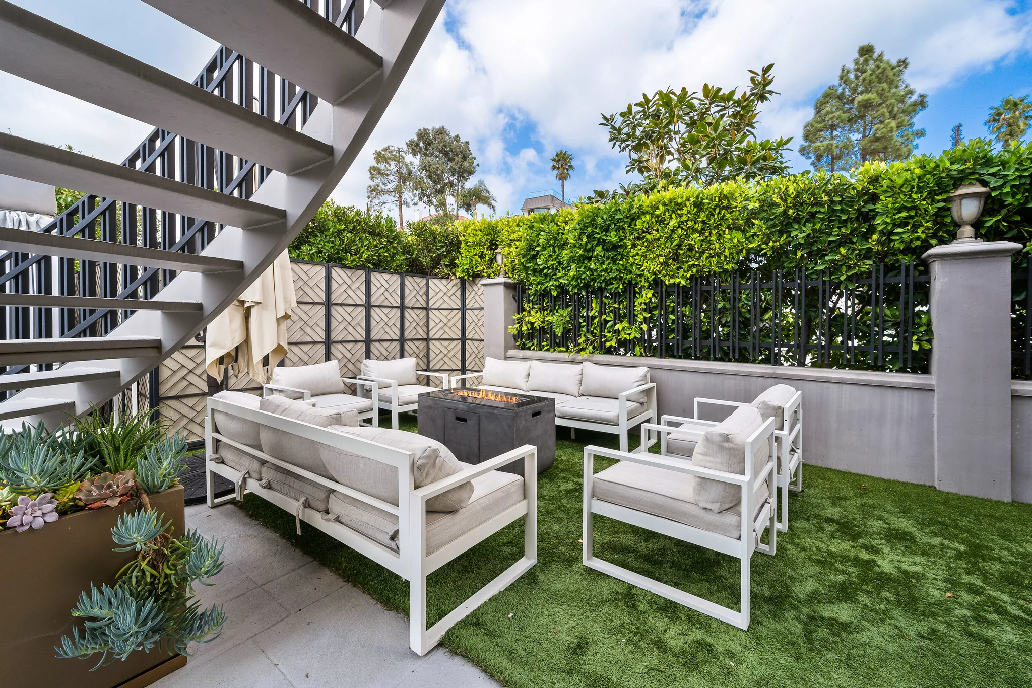 Fire-pit lounge on artificial lawn beneath the spiral stair, screened by a tall hedge for privacy