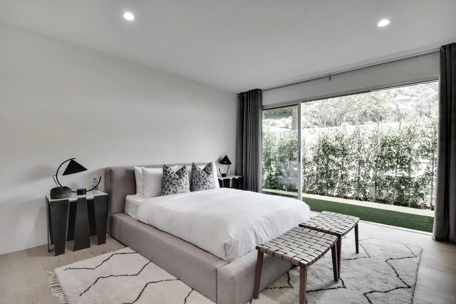 Ground-floor bedroom with upholstered bed, woven bench and sliding doors to a hedged lawn