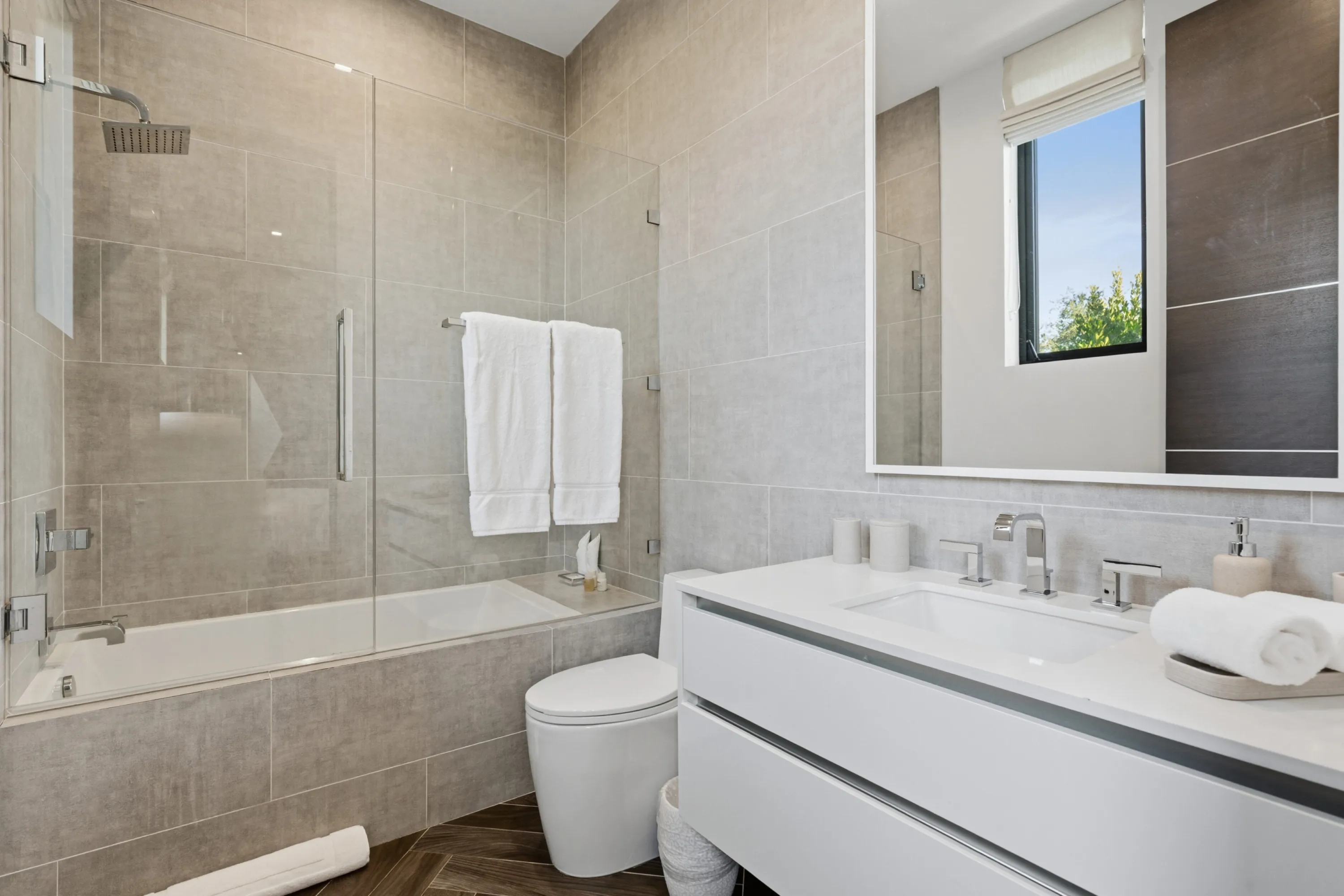 Guest bathroom with glass-screened tub-shower, stone tile and long white vanity beneath a daylit window