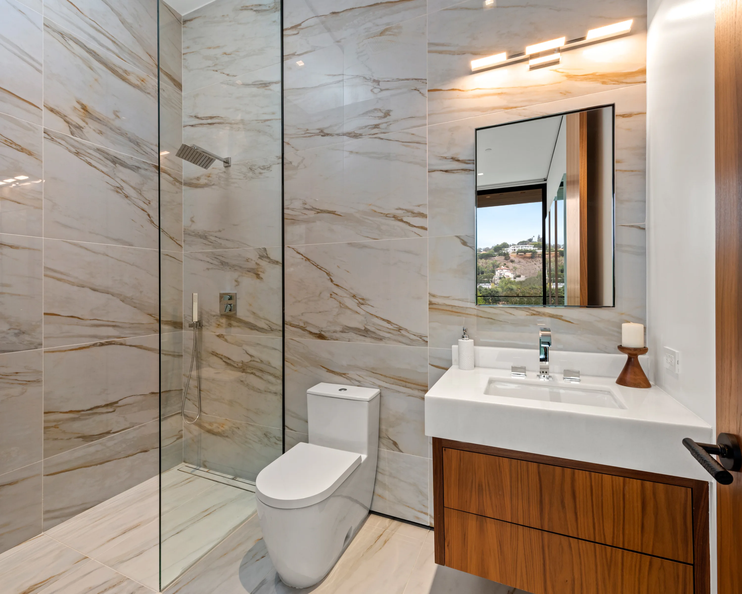 Compact guest bath in veined stone with walk-in rain shower and a hillside view caught in the mirror