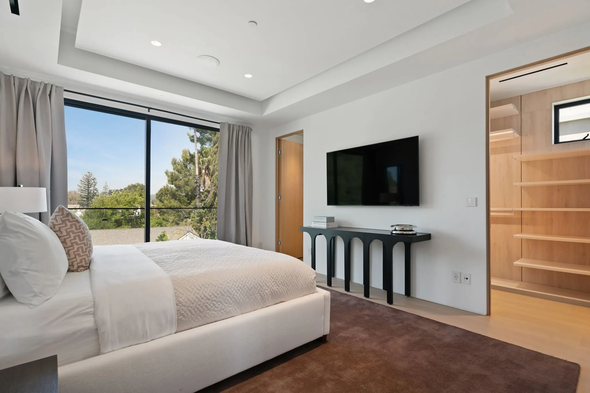 Bedroom opening to a treetop outlook through sliding glass, wall-mounted TV and open oak shelving closet