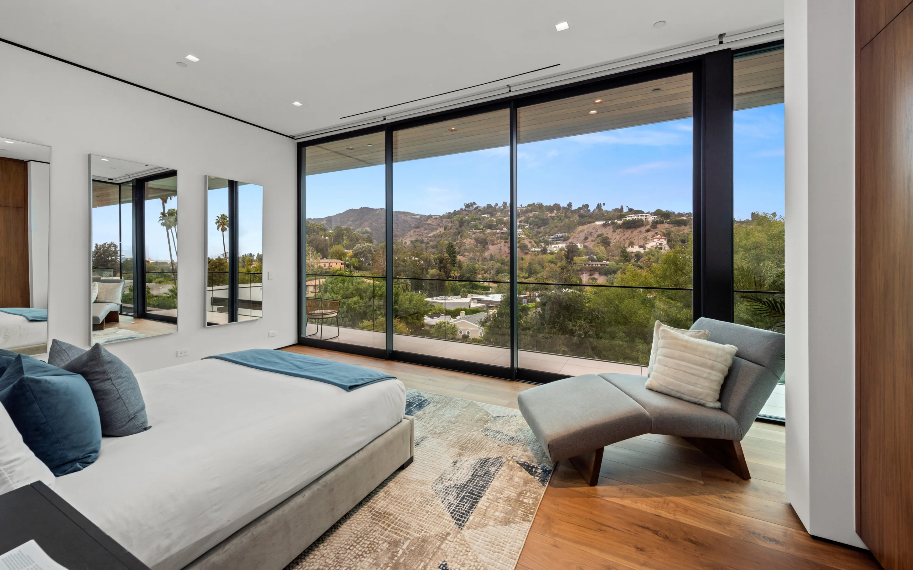 Corner bedroom framing canyon and hillside homes through full-height glass, curved lounge chair beside the bed