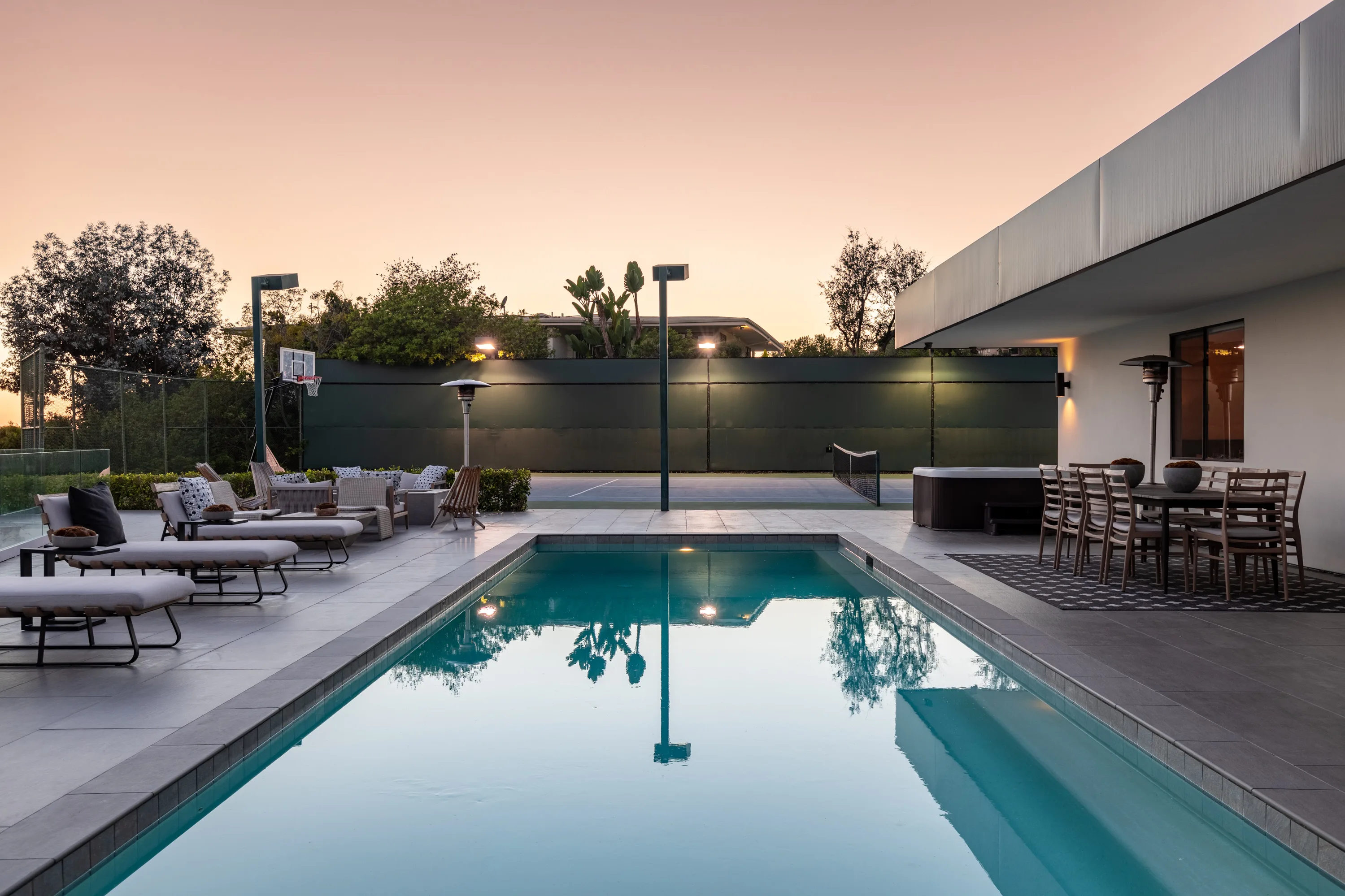 Pool glowing at dusk beside loungers, spa and outdoor dining under a peach sky