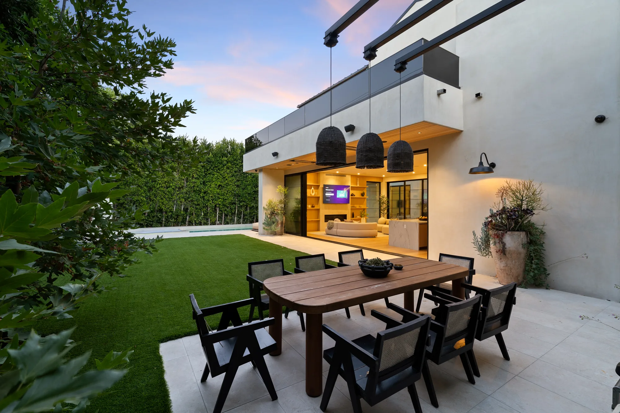 Dusk on the lawn terrace: outdoor dining table for eight under woven pendants, living room glowing behind open sliders