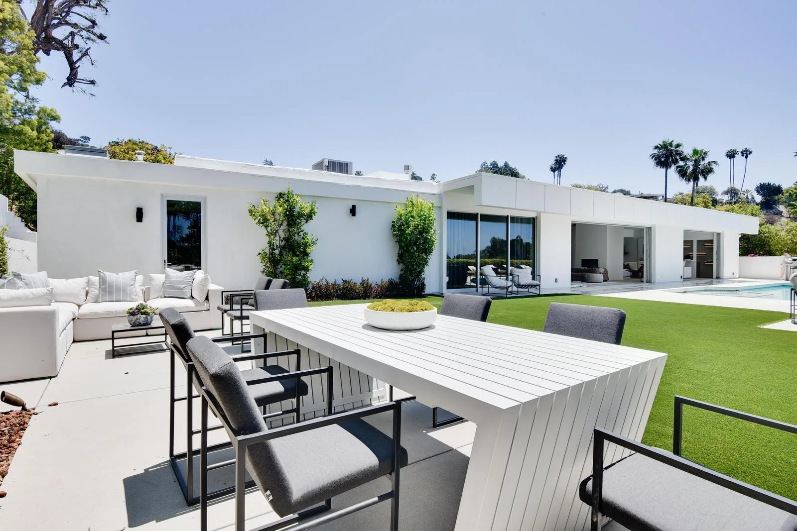 Poolside terrace with long white dining table and sectional sofa beside the lawn of this single-storey Beverly Hills estate