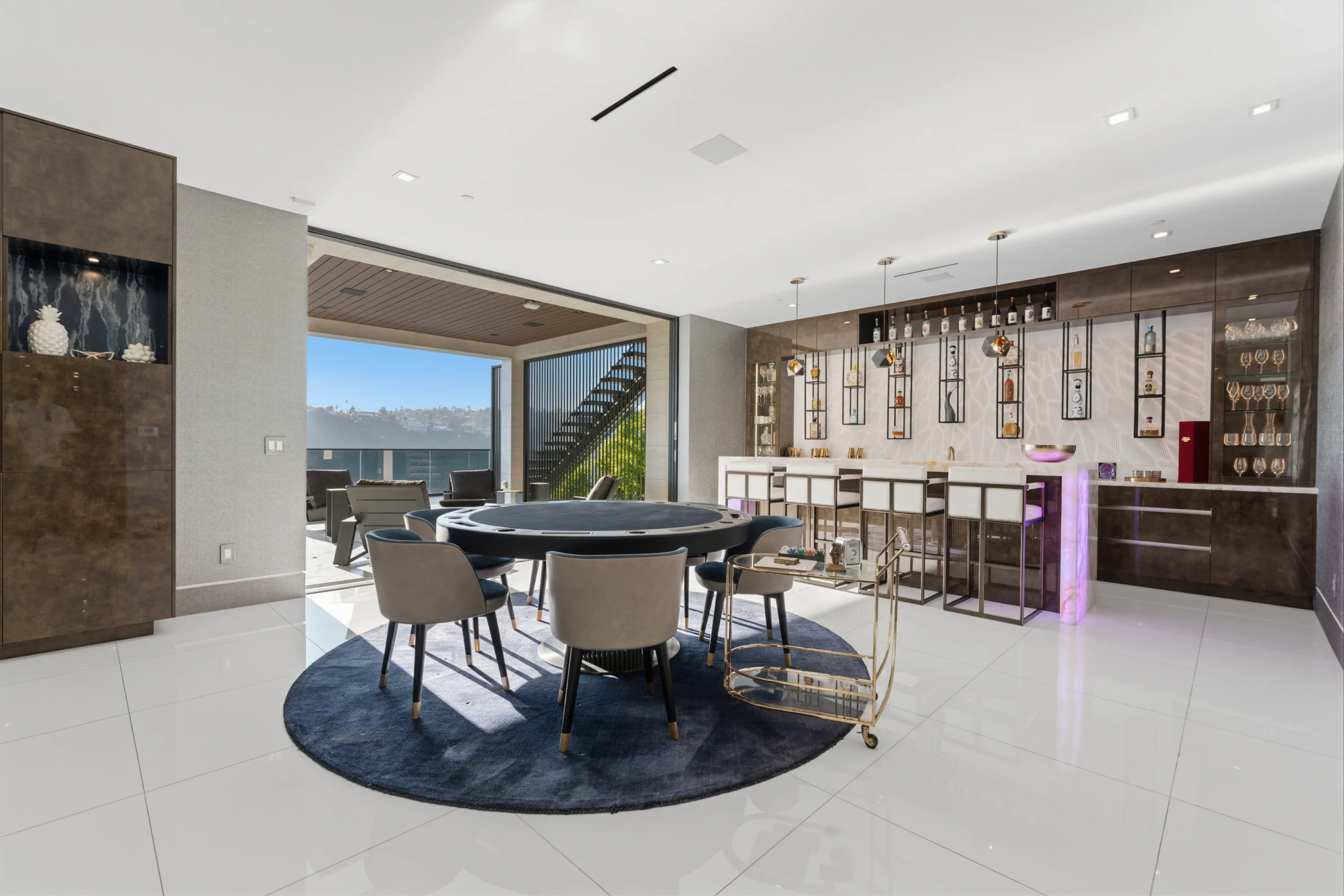 Poker table on a navy rug beside the backlit bar, sliding glass opening to the view terrace