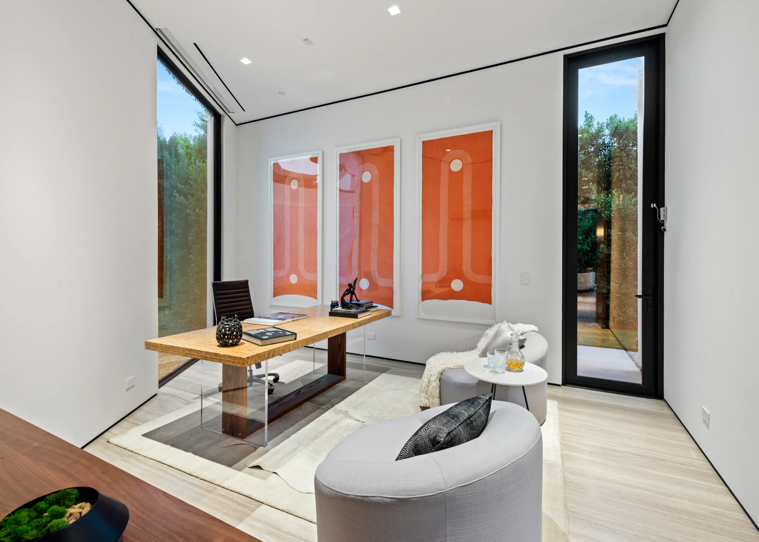 Home office with a floating desk, triptych of orange artworks and glass doors to a hedge-lined courtyard