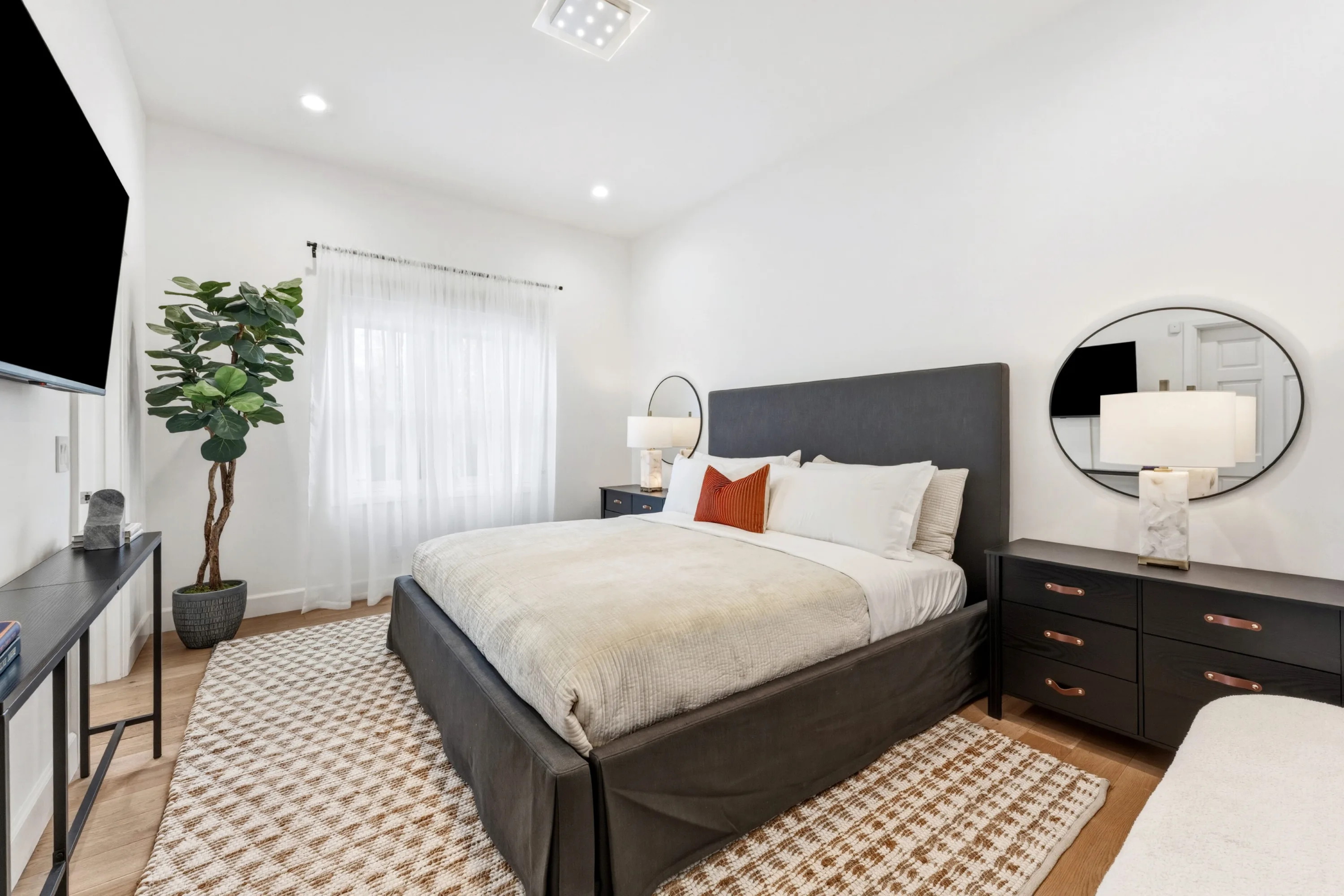 Guest bedroom with charcoal upholstered bed, round mirror, wall-mounted TV and sheer-curtained window
