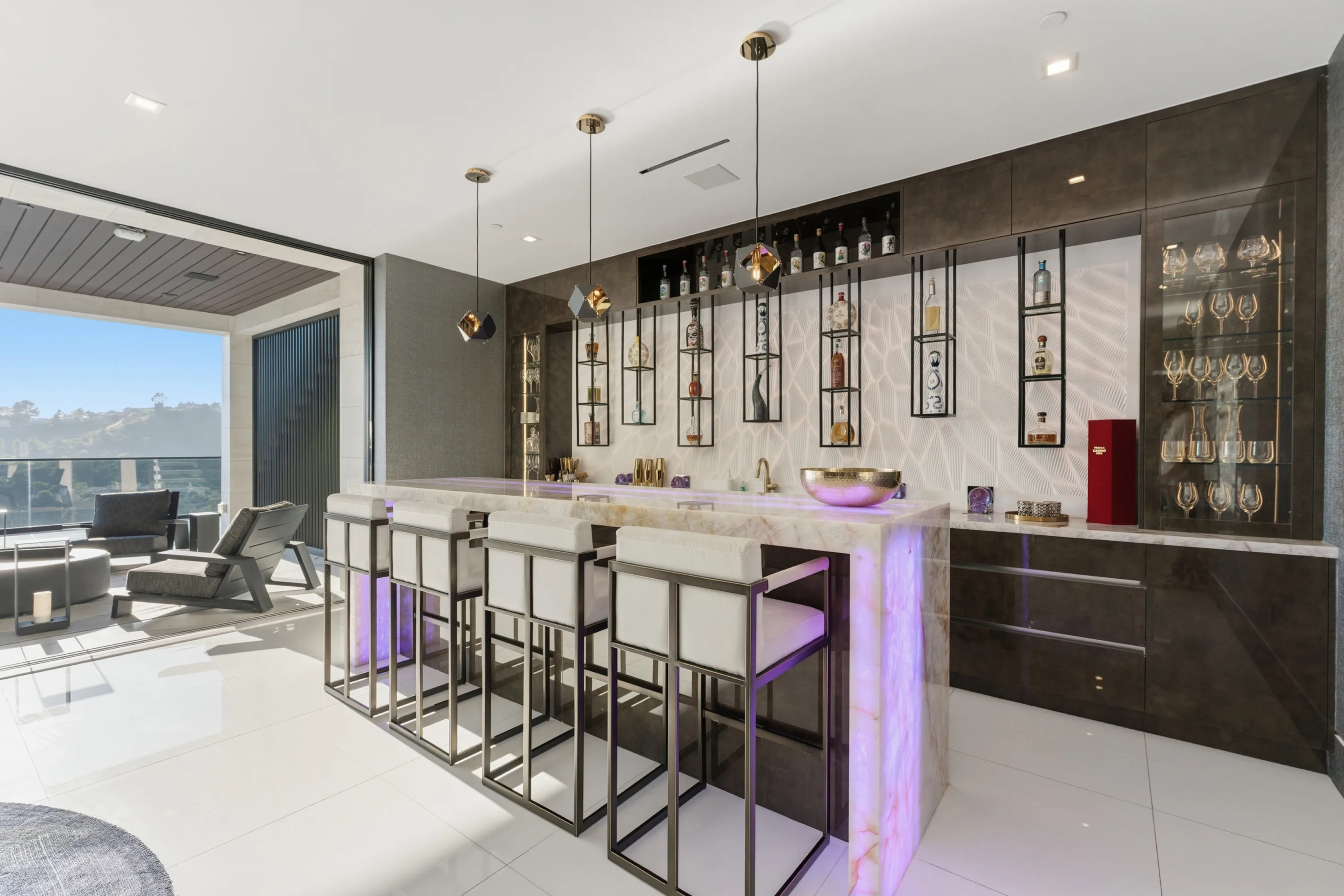 Backlit onyx bar counter with four stools, spirits displayed on floating shelves and terrace beyond