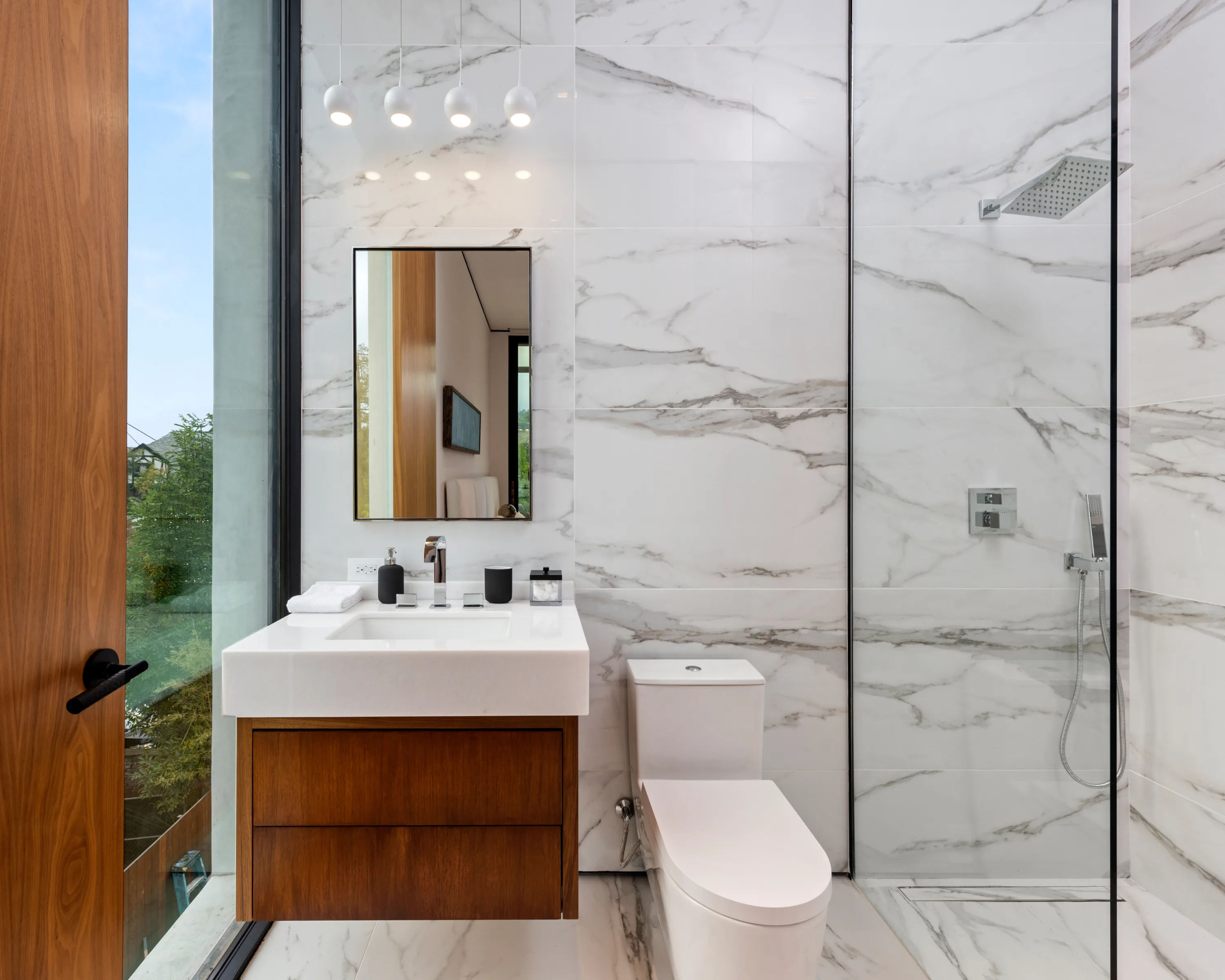 Slim guest bath with pendant lights, walnut vanity and a glass shower beside a window onto the treetops