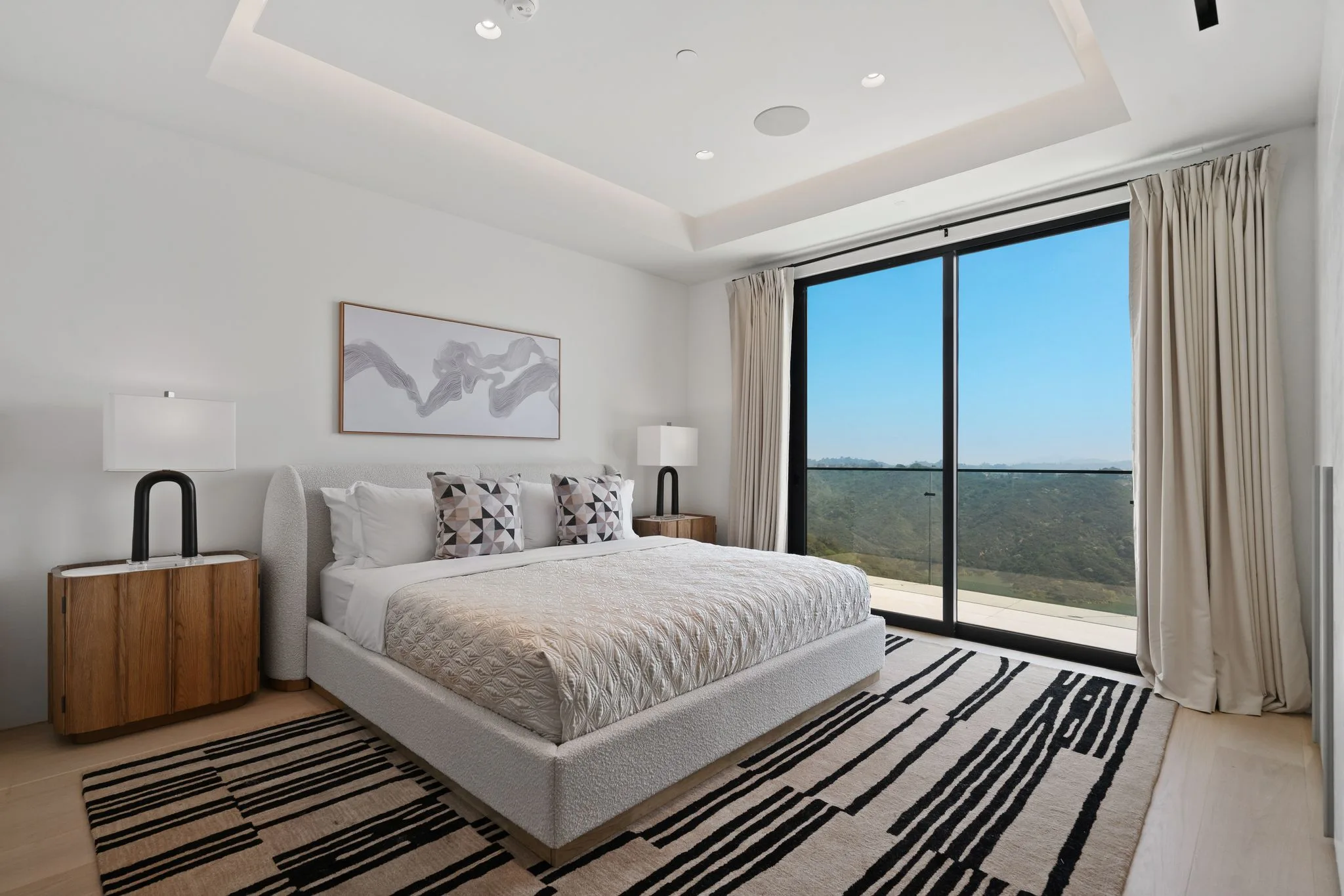 Boucle-upholstered bed facing a full-height slider onto the green canyon, graphic striped rug underfoot