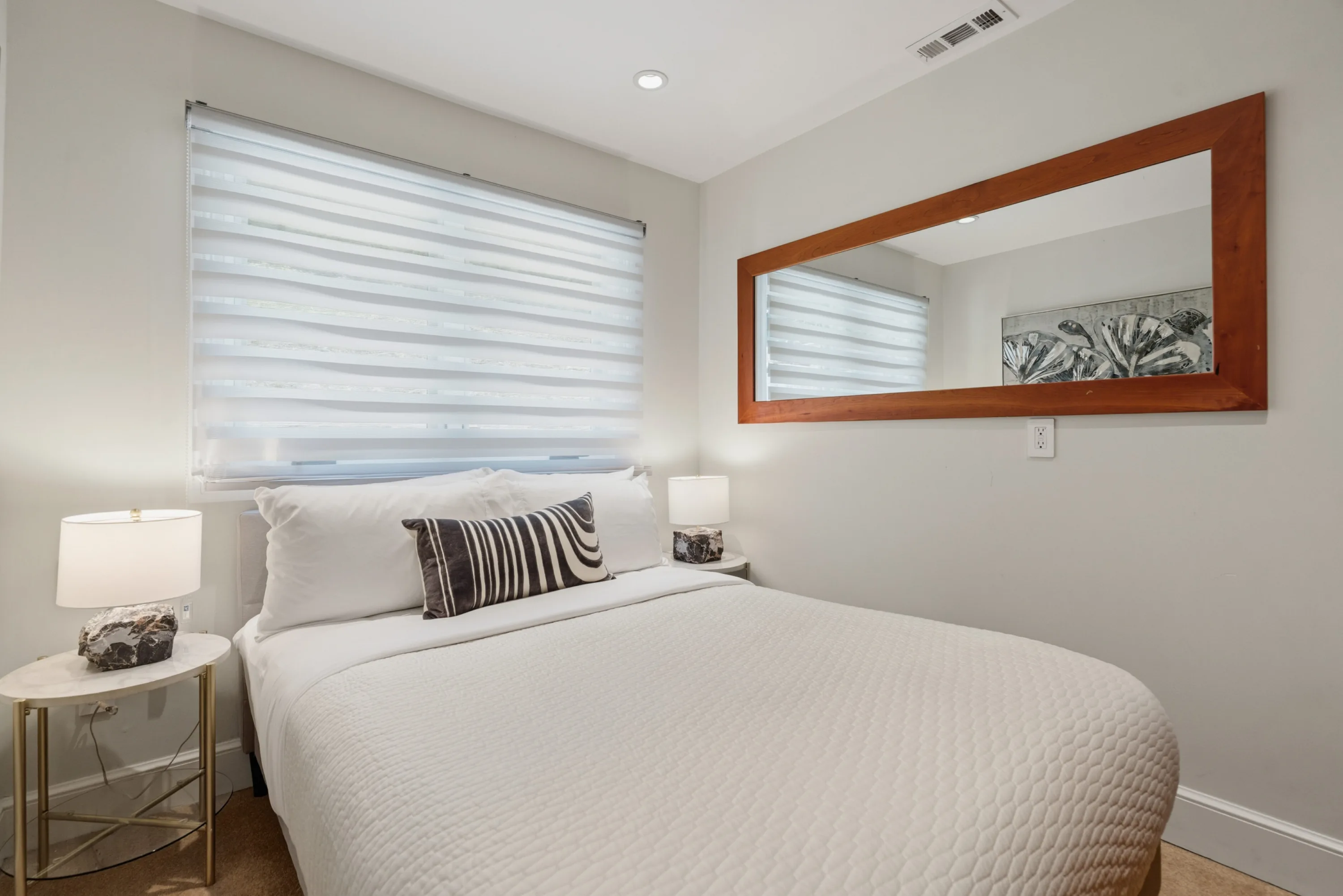 Compact guest room with crisp white bedding, softly shaded window and a long cherrywood mirror above the bed