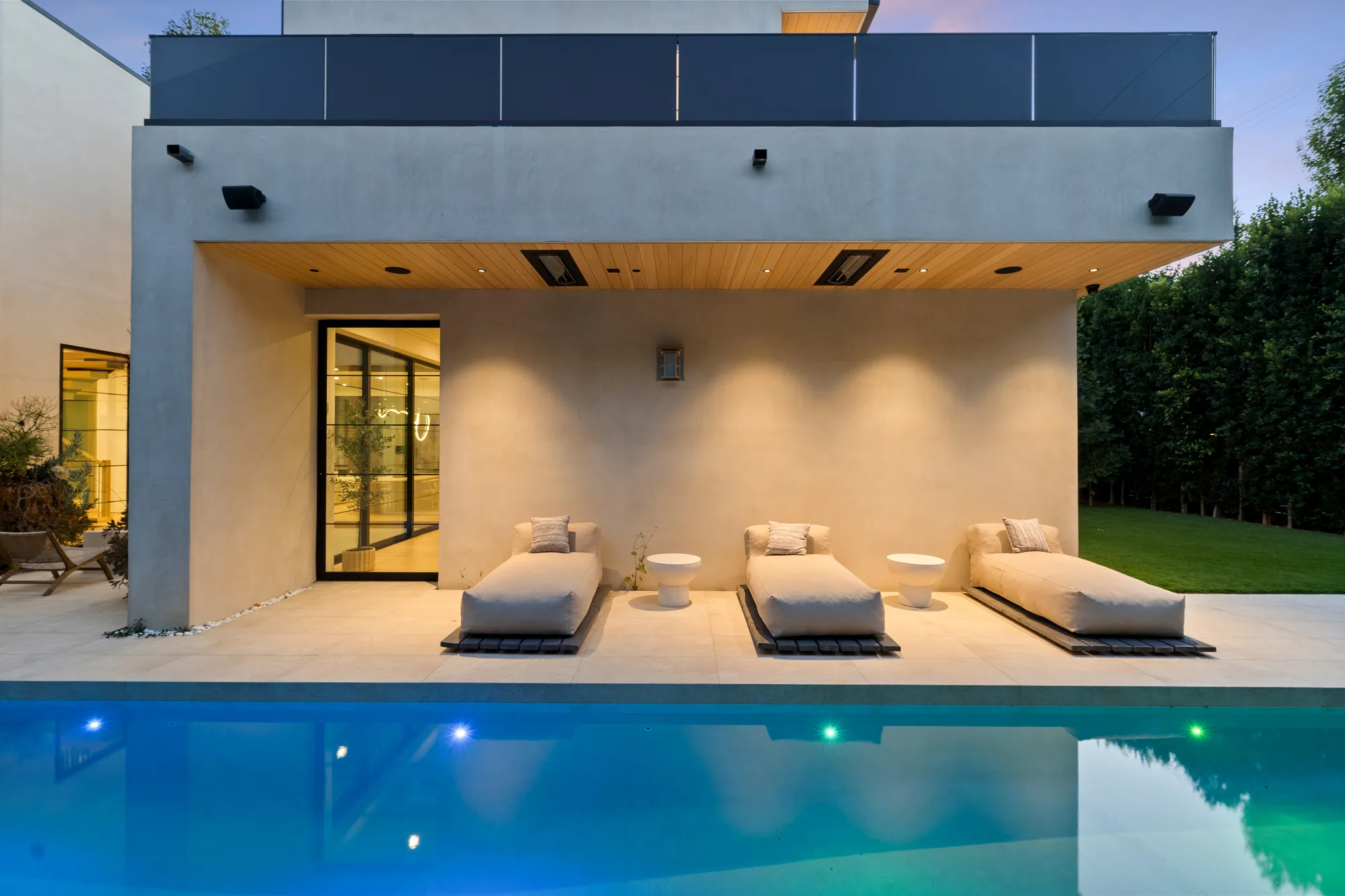Three poolside daybeds under the heated loggia, pool lights glowing blue at blue hour