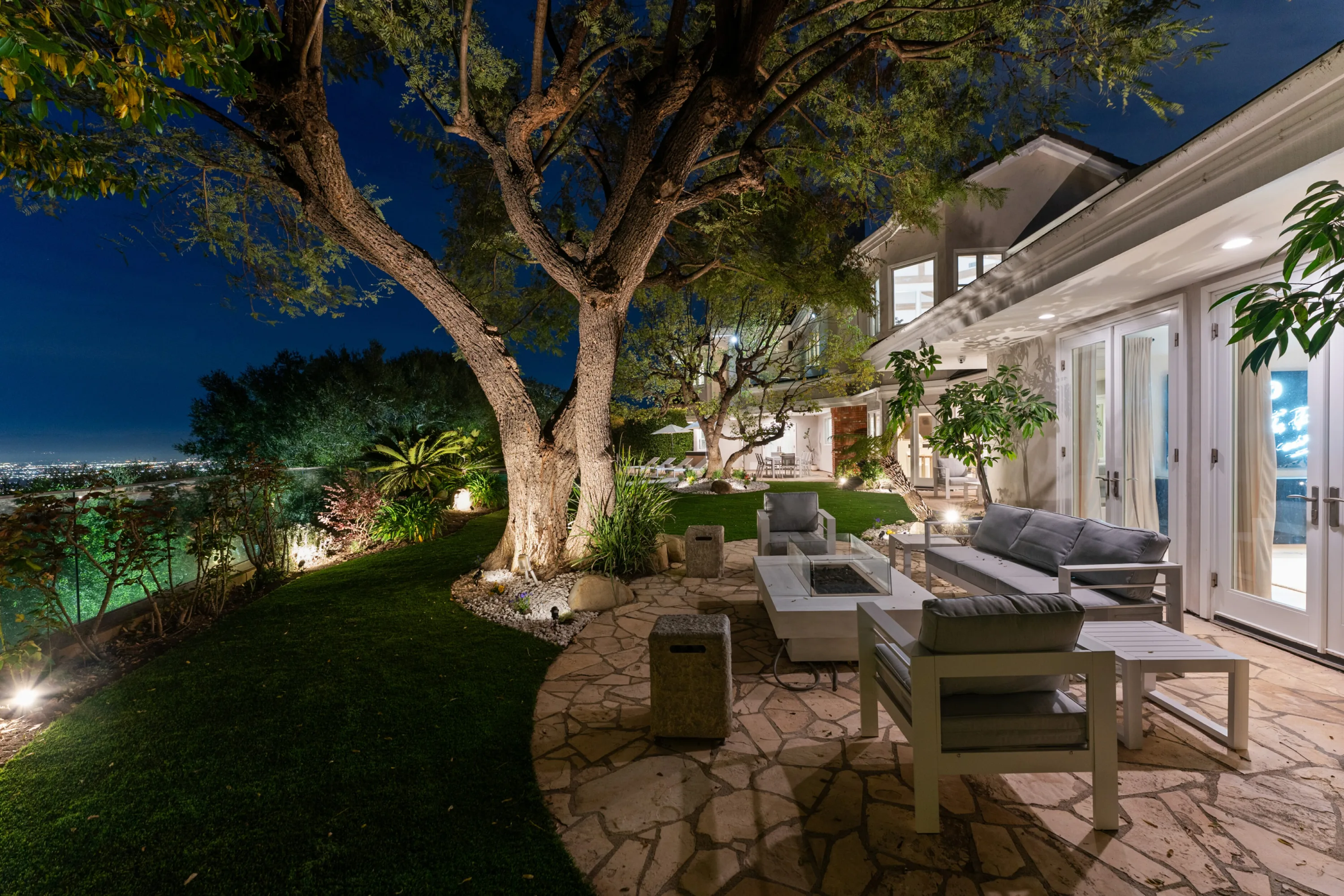 Flagstone terrace with fire-table lounge under a lit oak at dusk, city lights glittering beyond the lawn