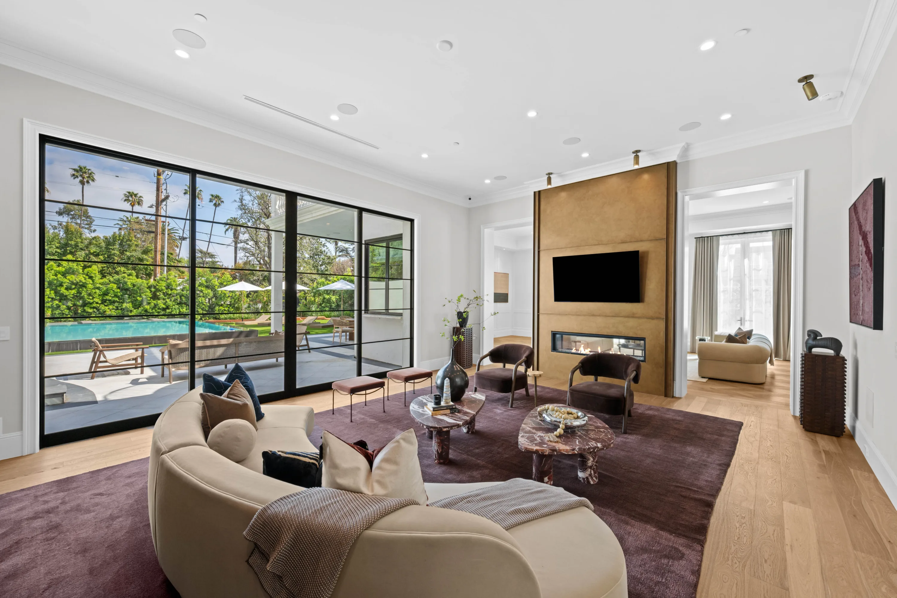 Great room with curved sofa, linear fireplace below the TV and steel-framed sliders opening to the pool terrace