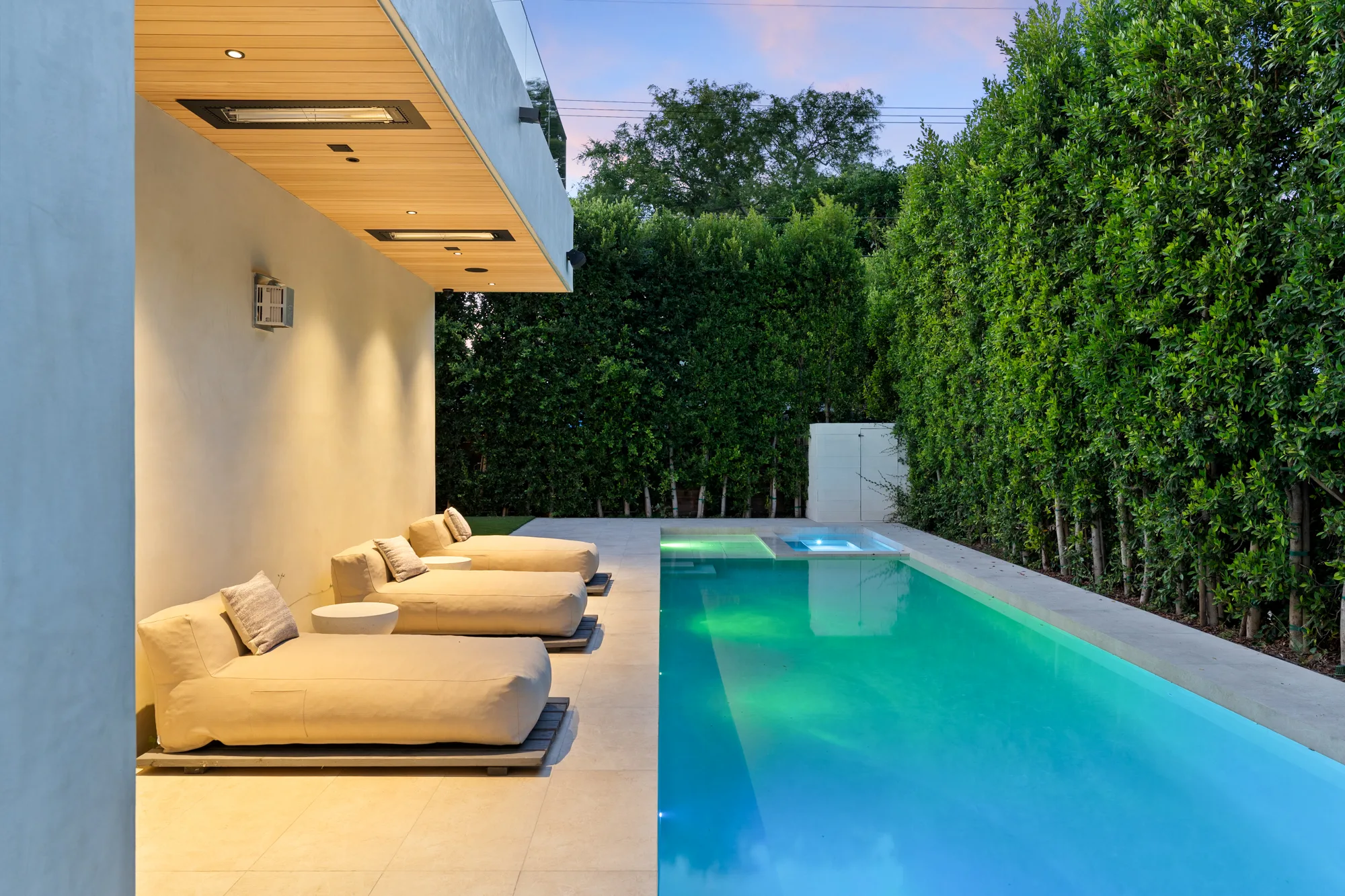 Lap pool and spa lit green and blue at dusk, cushioned sun beds lined along the covered terrace