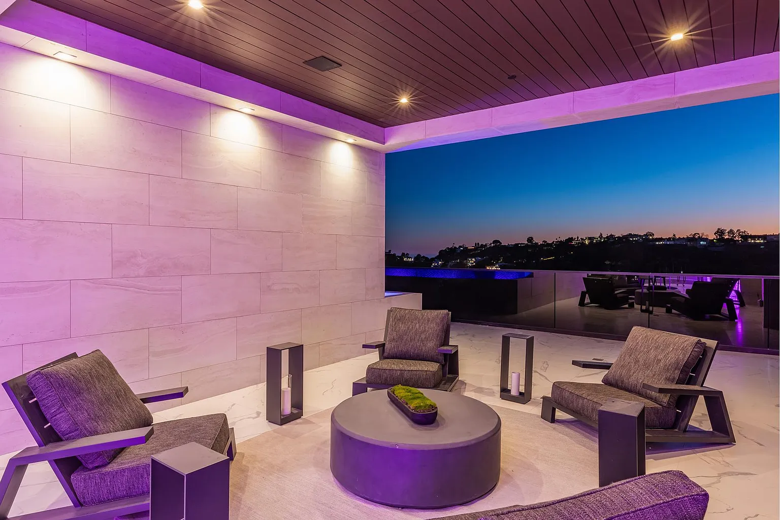 Covered terrace lounge under violet cove lighting at dusk, city lights and pool glowing beyond