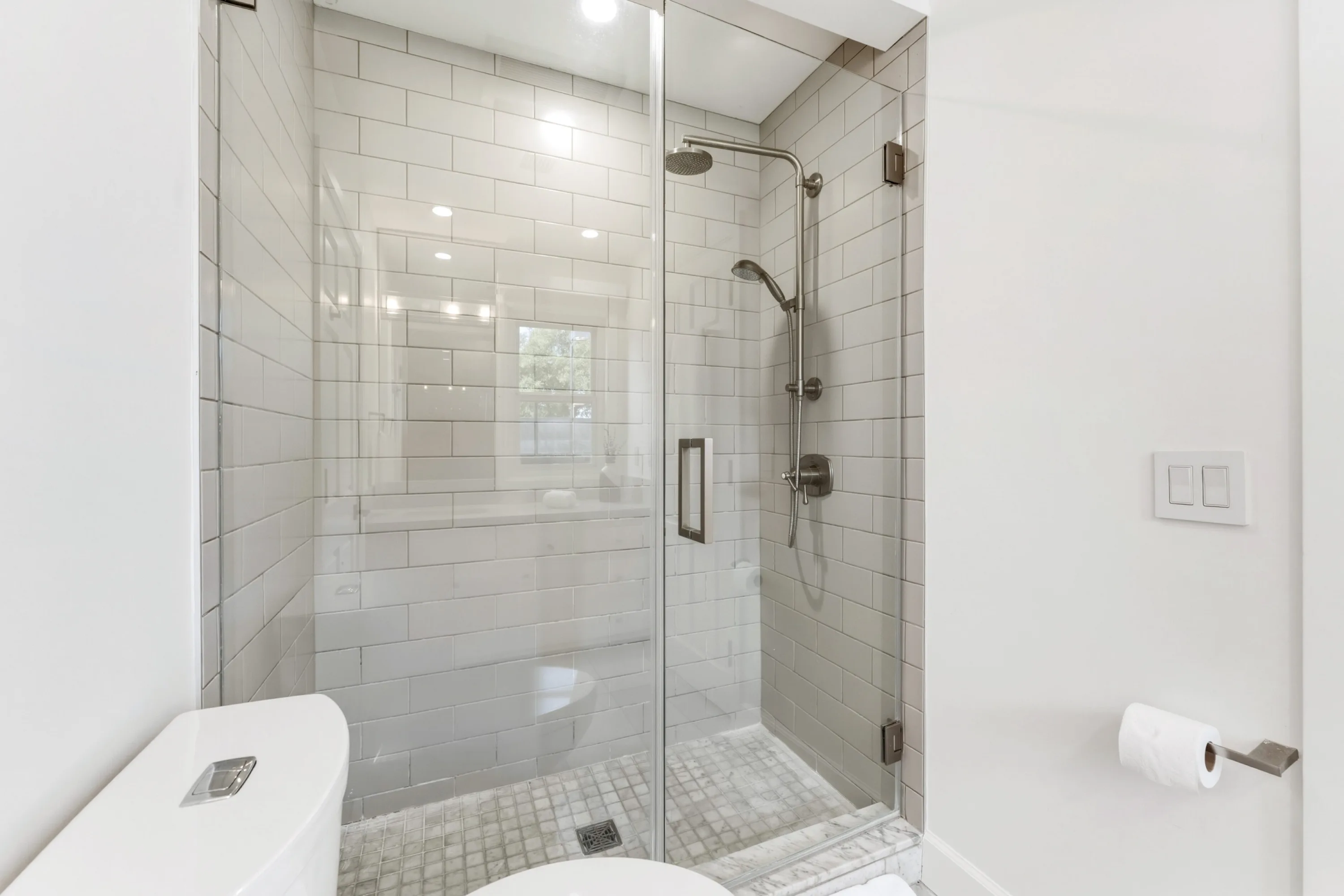 Walk-in glass shower tiled in pale subway tile with rainhead and mosaic marble floor