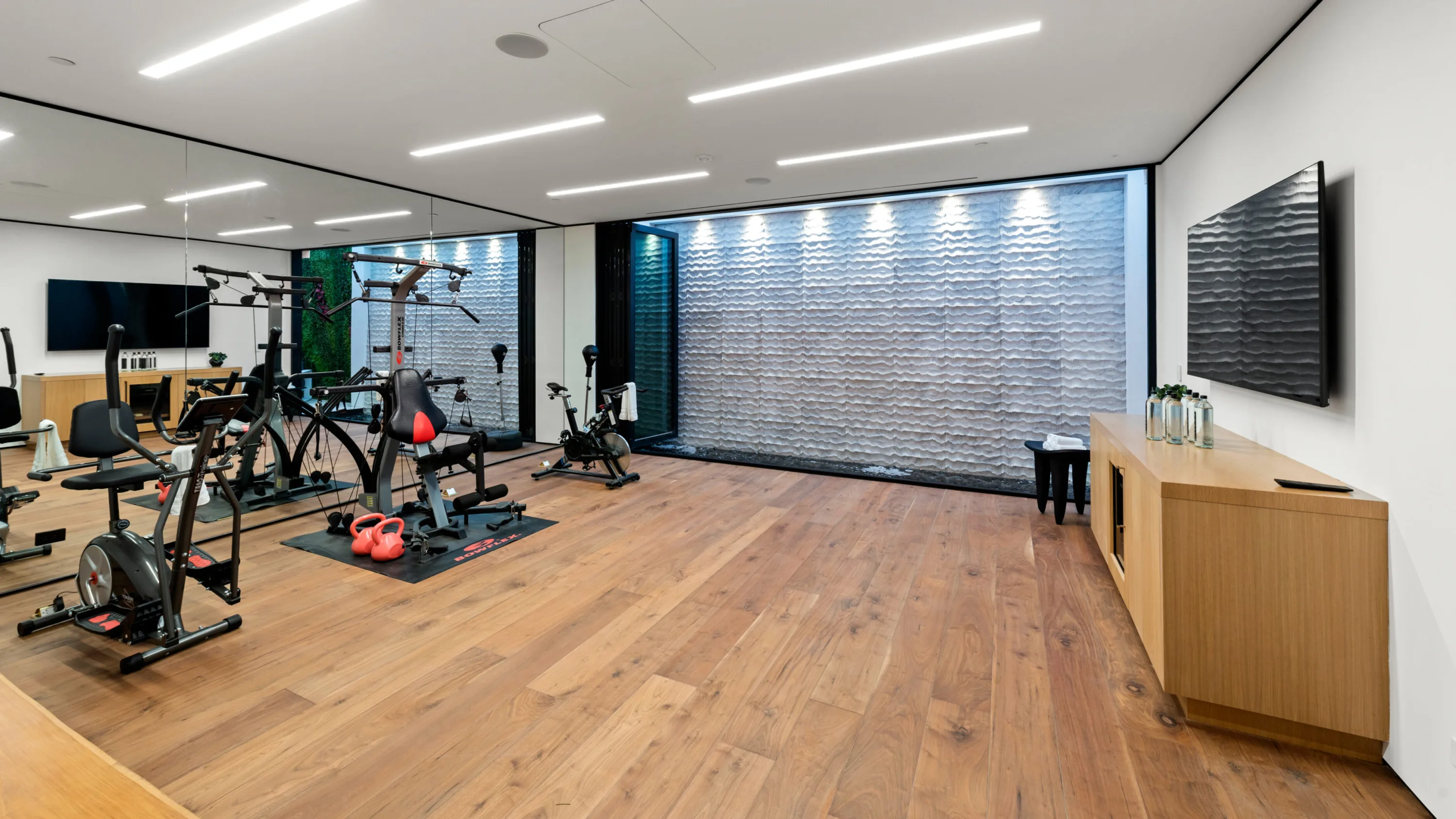 Mirrored gym with cycles, cable machine and kettlebells facing a backlit textured stone water wall