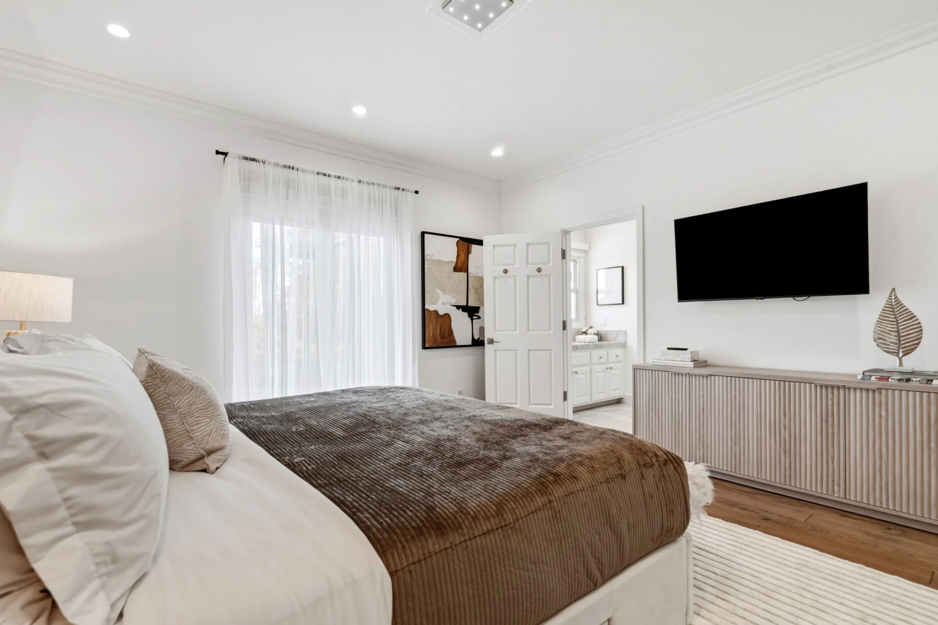 Bedroom looking toward its ensuite, with fluted credenza, mounted TV and sheer-draped window