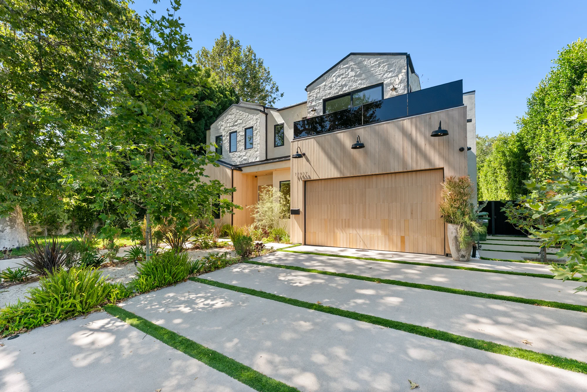 Street frontage of the Valley Village house, pale timber garage and stone gables above a ribbon-grass driveway