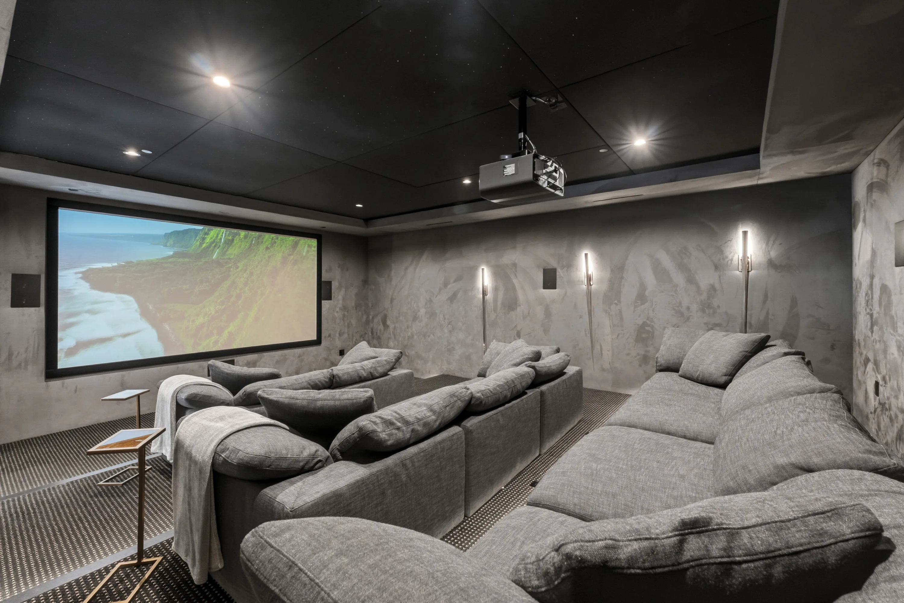 Home cinema with tiered grey sectional seating, ceiling projector and coastal footage on the wide screen