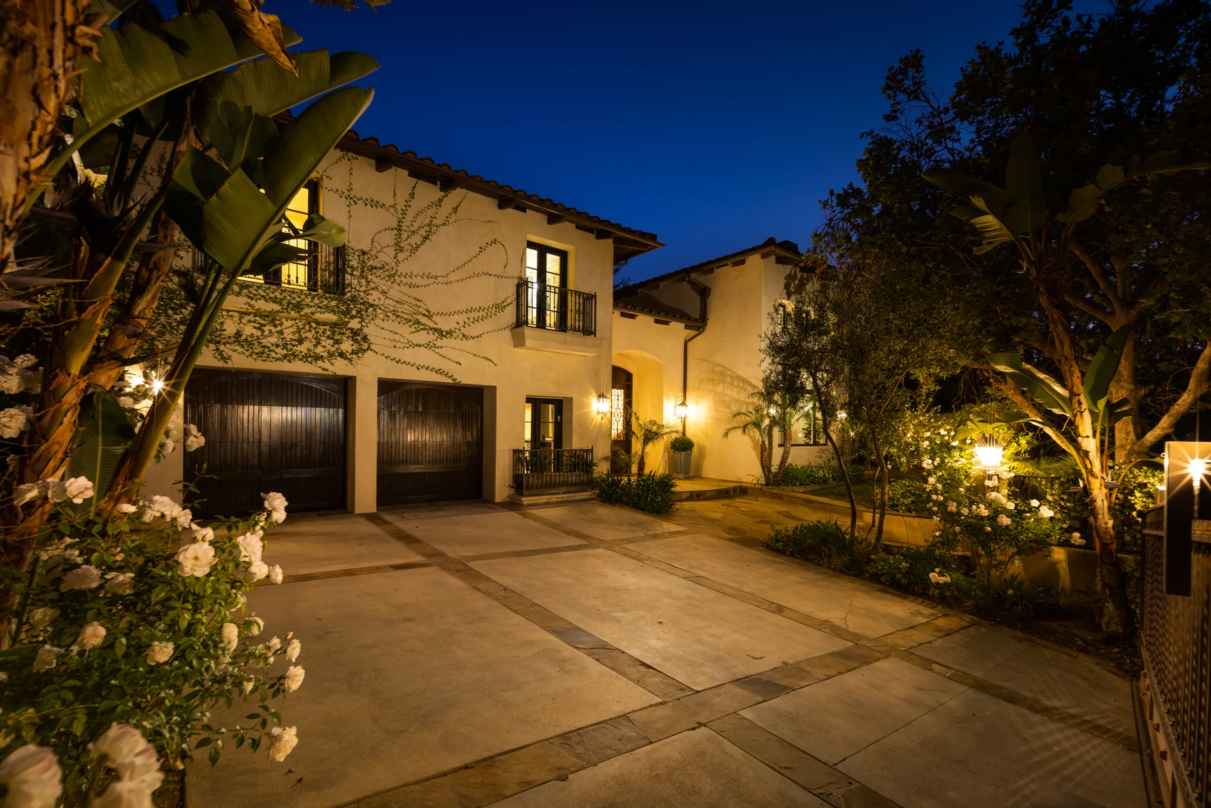 Spanish-style facade and stone motor court lit at dusk, framed by roses and banana palms in Beverly Hills