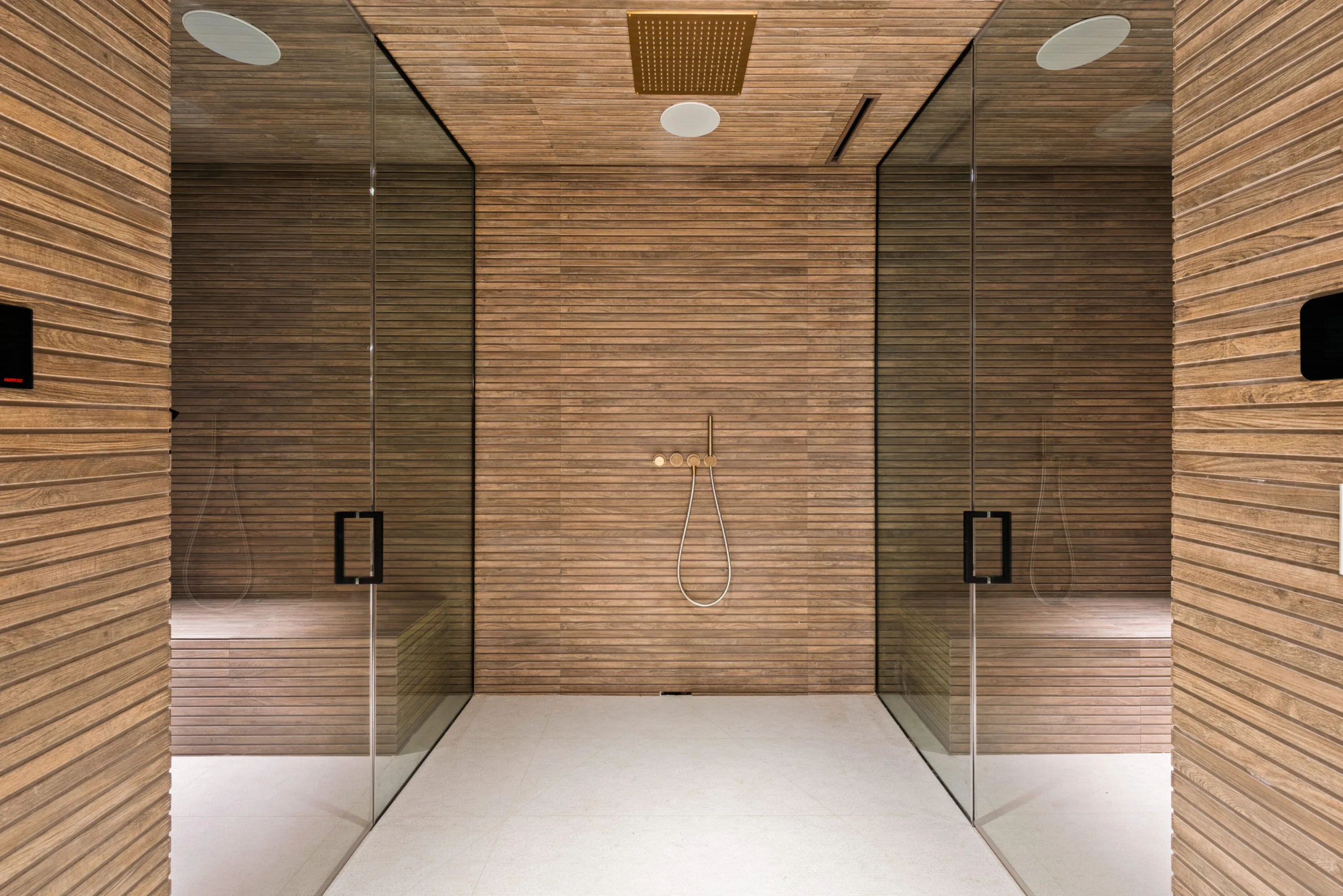 Twin glass steam rooms in slatted teak flanking a central shower with a brass ceiling rain head