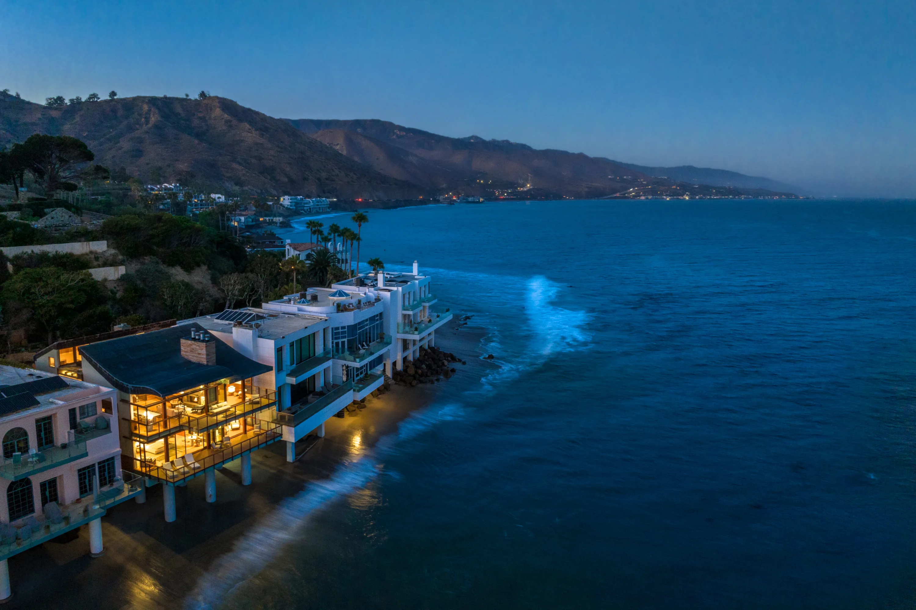 Malibu oceanfront home on pilings, its lit interiors glowing over the surf as the coastline fades into dusk