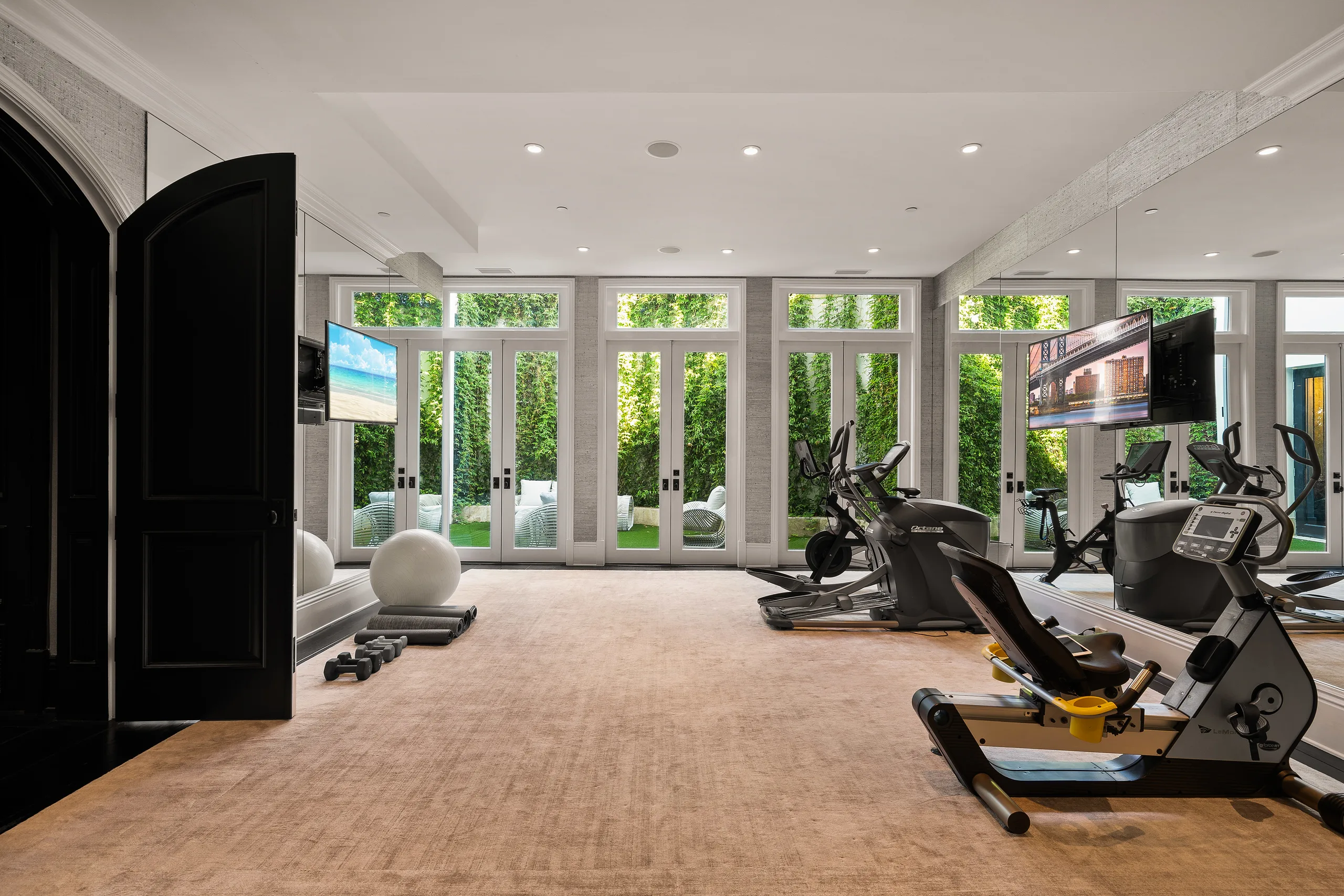 Mirrored home gym with cardio machines, free weights and wall-mounted screens facing ivy-framed French doors