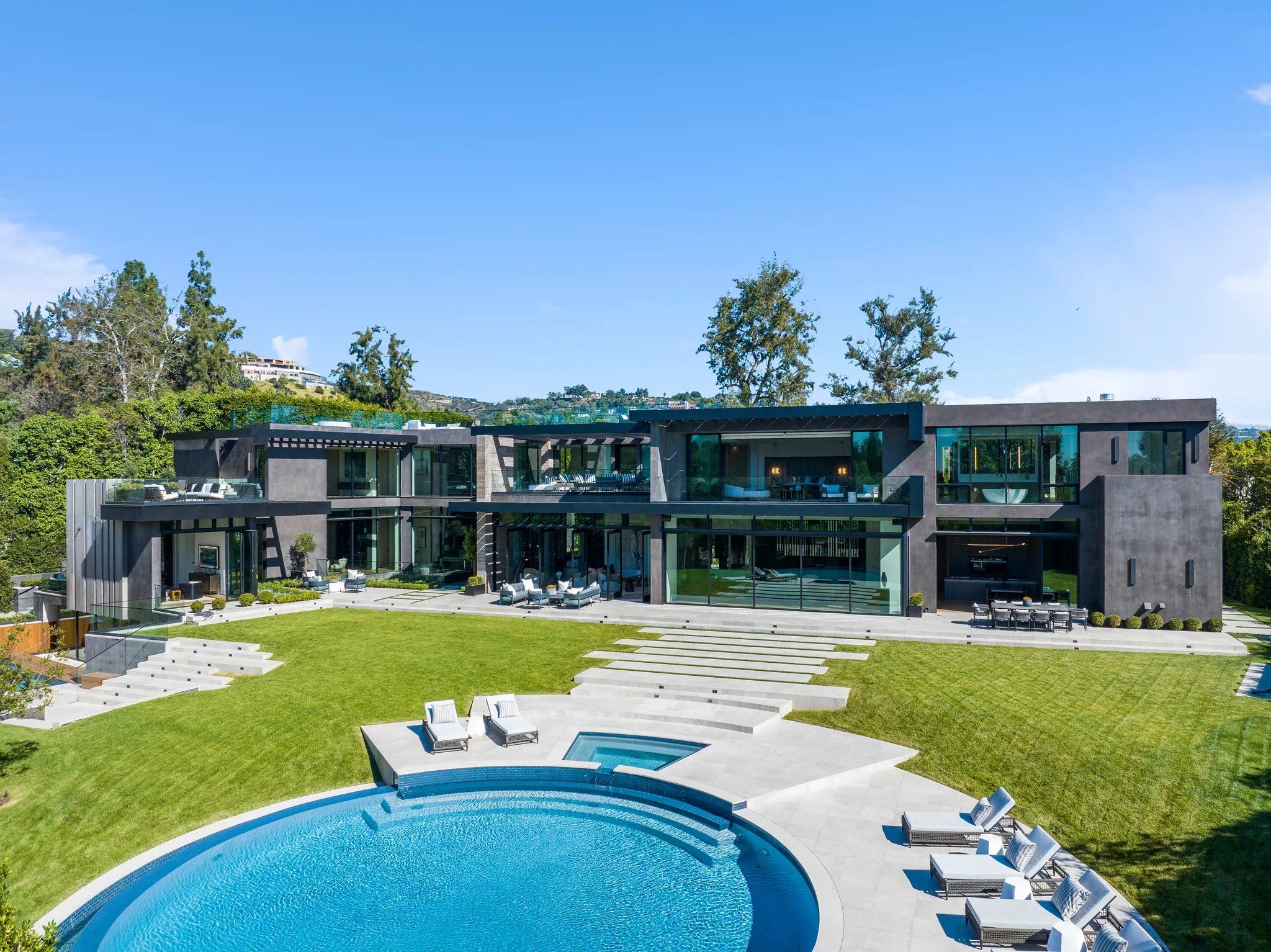 Glass-walled contemporary residence opening onto a sweeping lawn, stone steps and circular pool with sun loungers