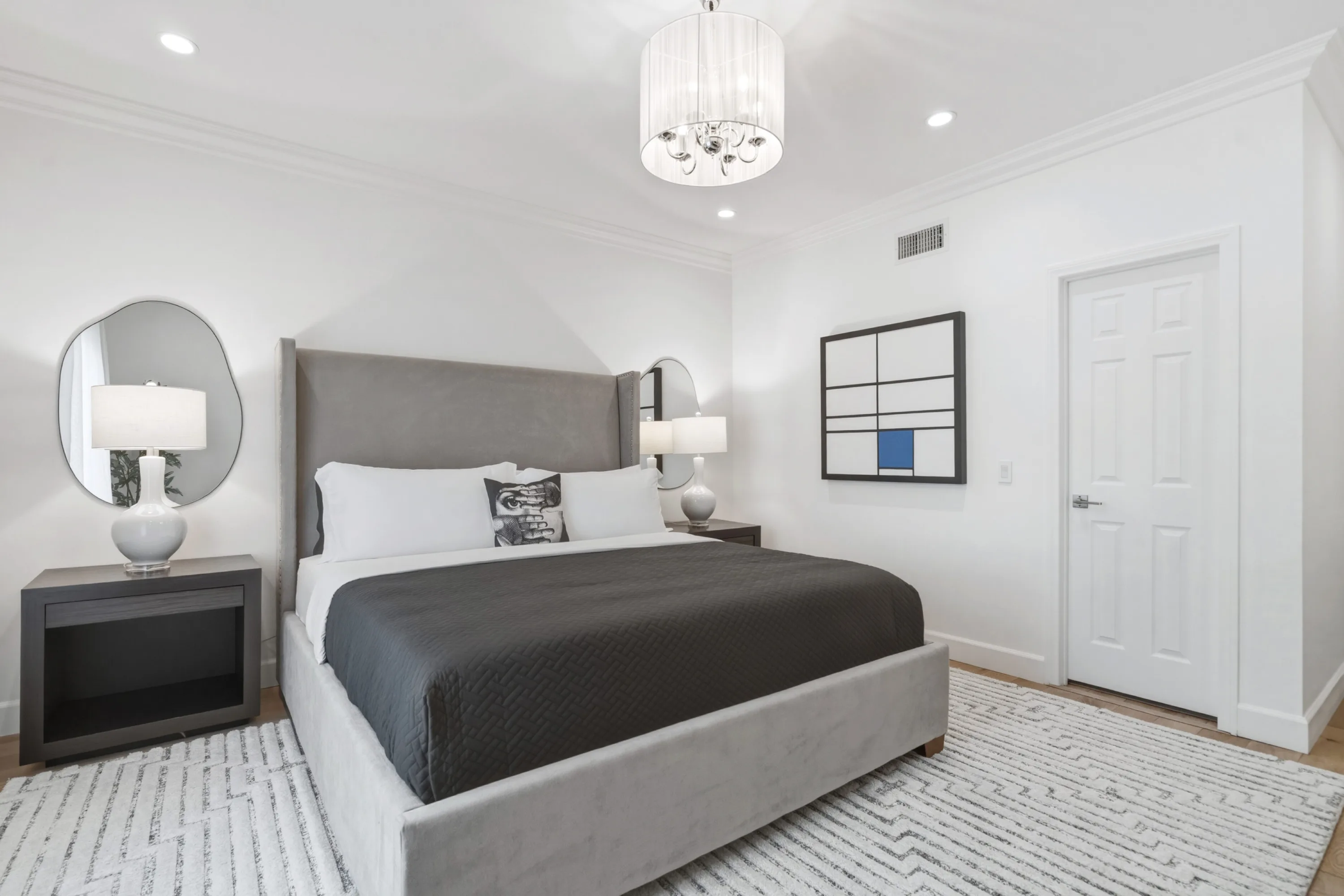 Grey velvet king bed beneath a drum chandelier, flanked by organic-shaped mirrors