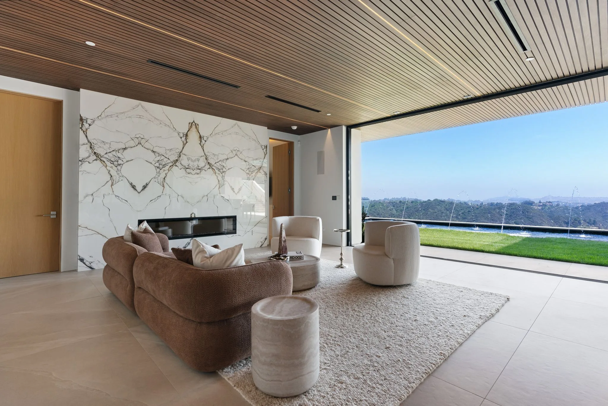 Sitting room with its glass wall retracted, opening onto lawn and hazy canyon beyond the fire feature