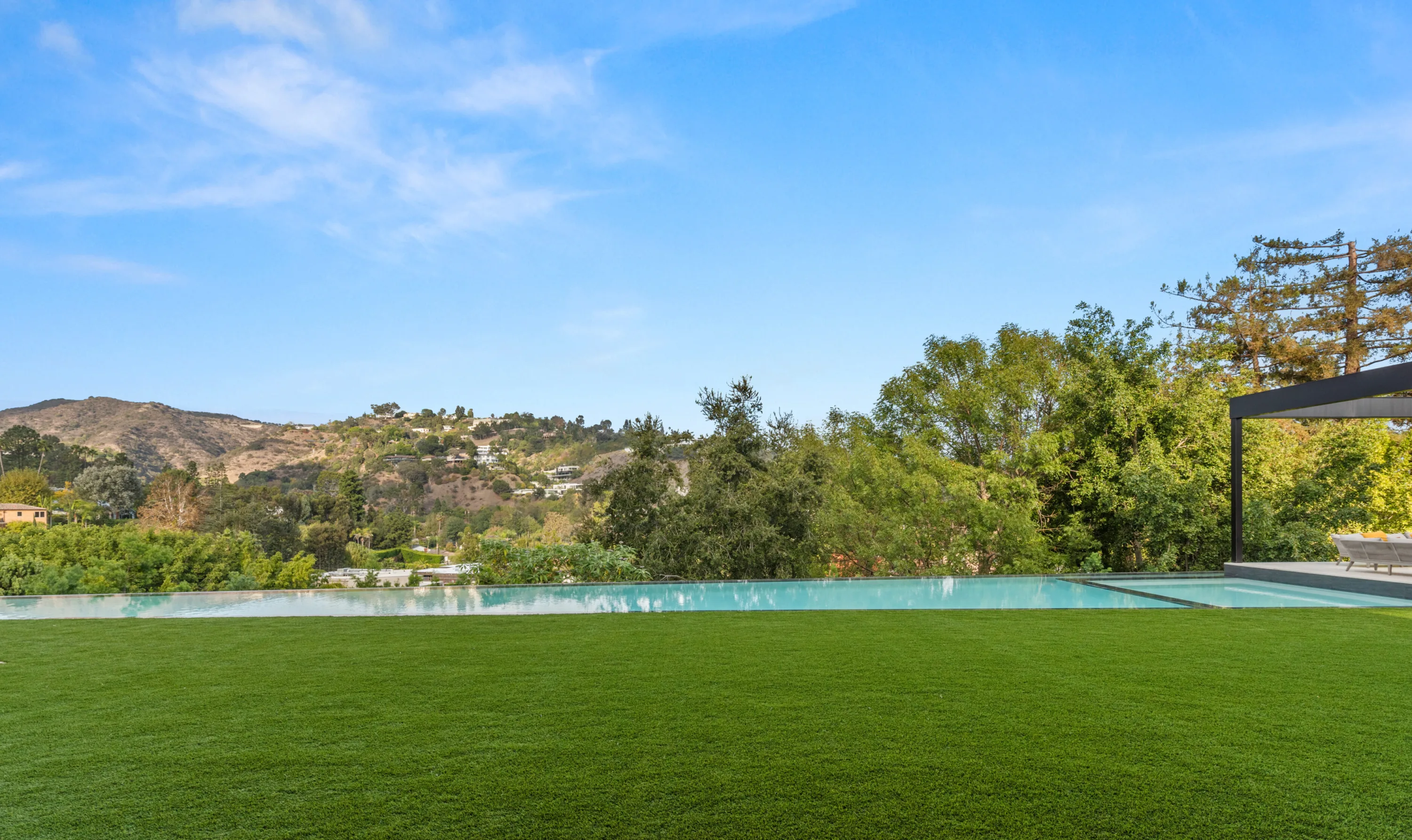 Zero-edge pool meeting a broad lawn, hillside homes and treetops filling the horizon