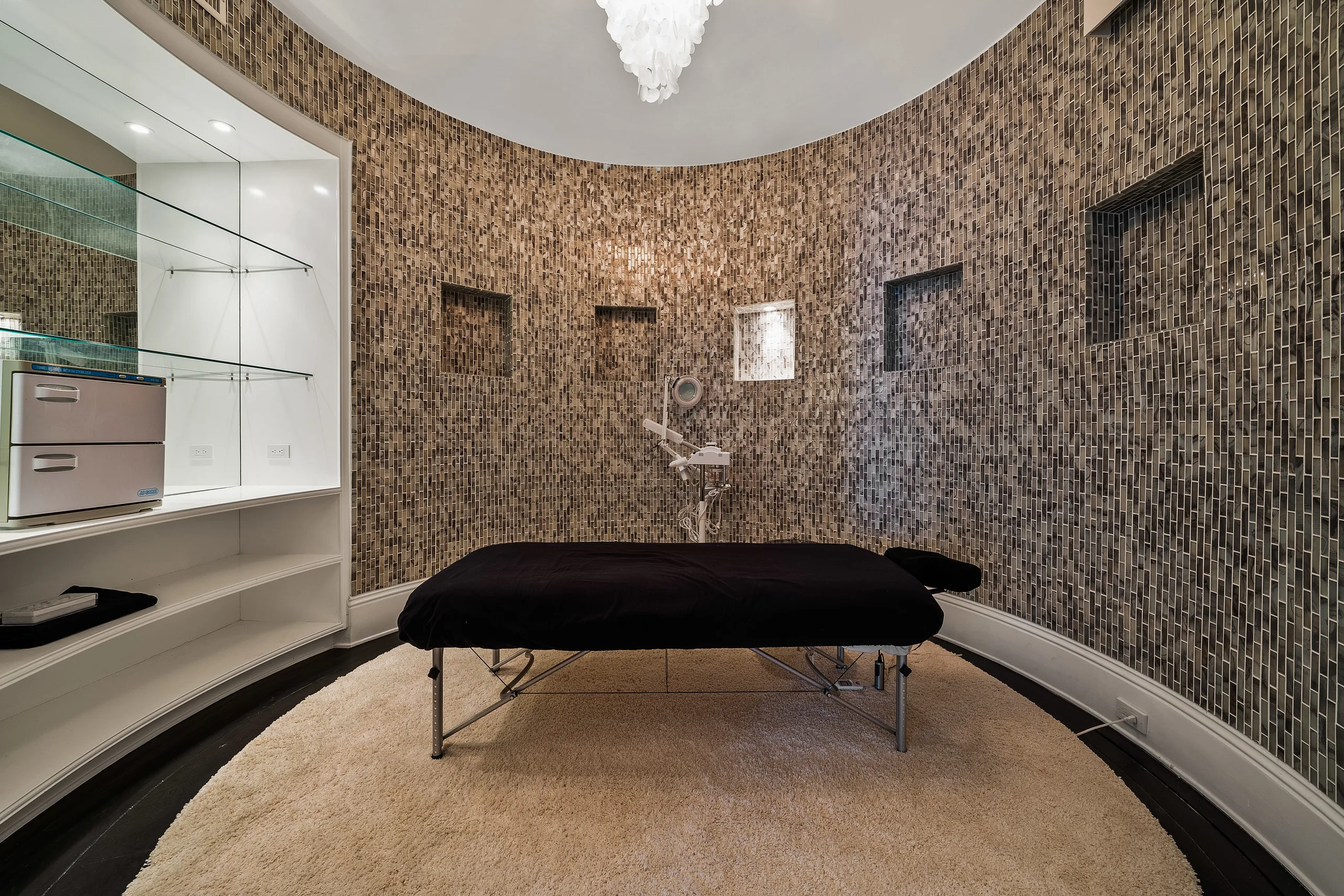 Curved mosaic-tiled treatment room with massage table, towel warmer and deep pile rug for in-villa spa days