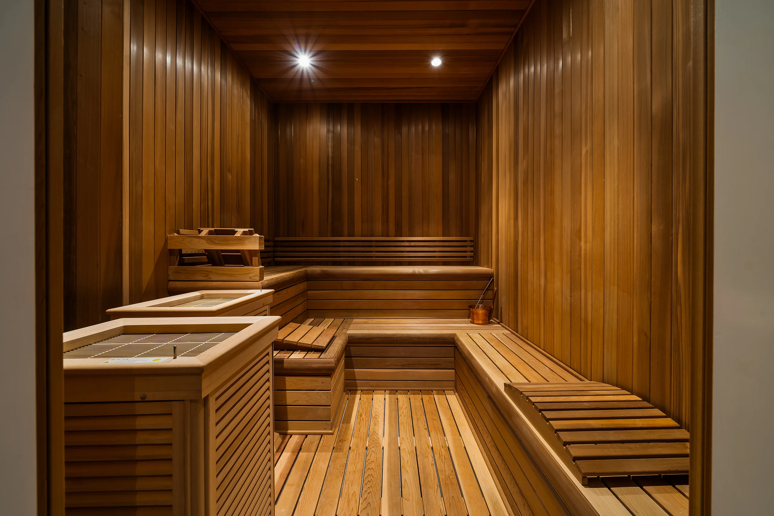 Cedar-lined sauna with wraparound benches, slatted floor and a copper ladle beside the heater