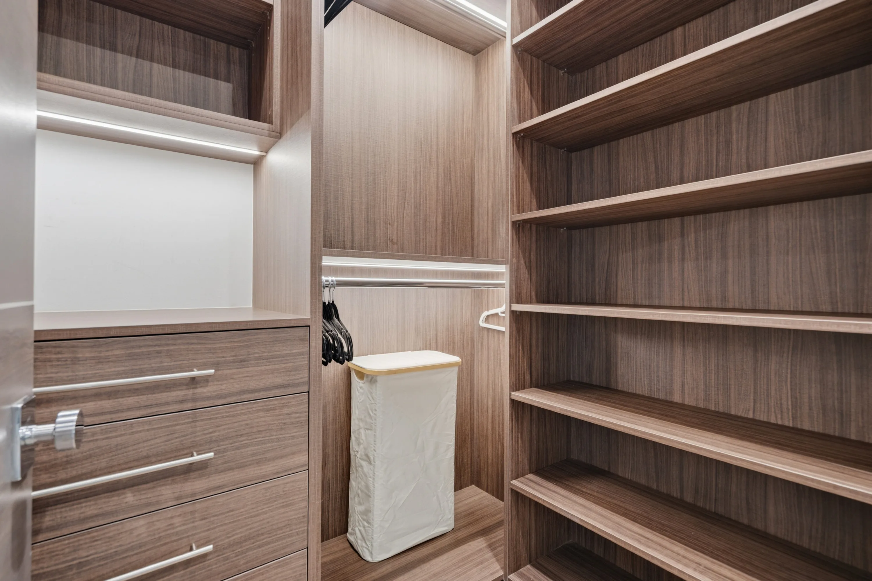 Walk-in wardrobe fitted in warm walnut with lit shelving, drawer bank and hanging rail