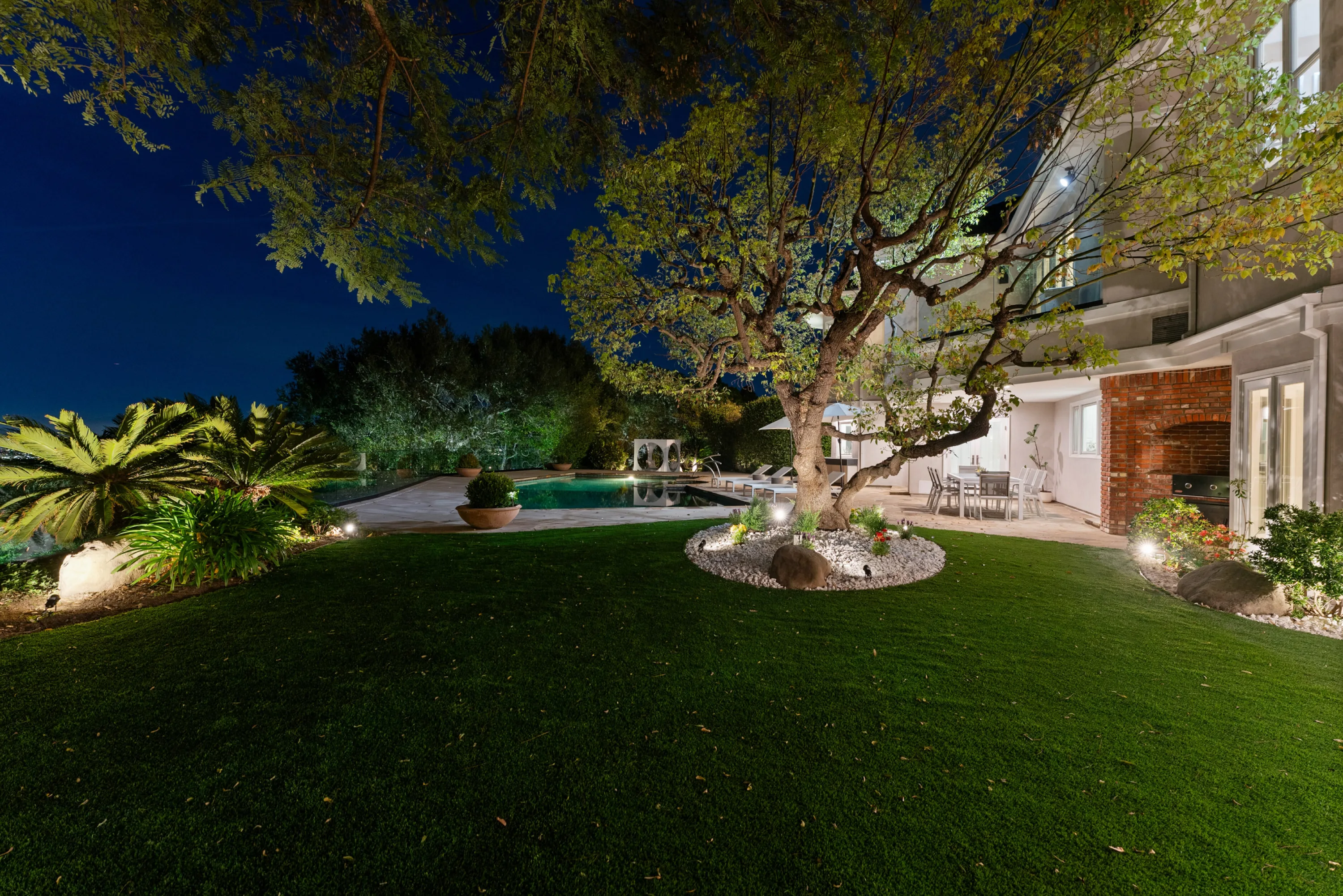 Sweeping lawn and uplit pool at blue hour, with a brick barbecue and outdoor dining set against the house