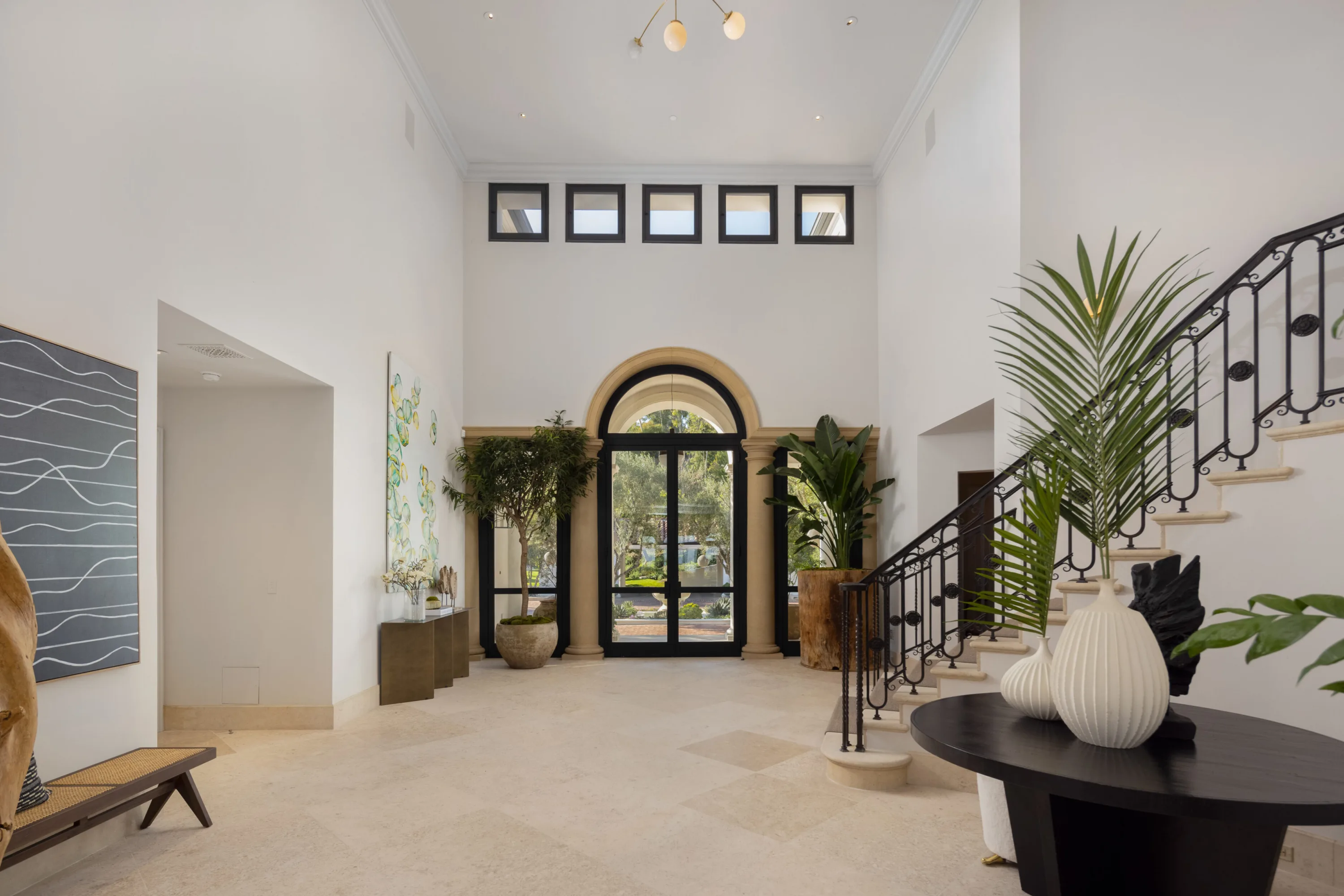 Double-height entry hall with arched glass doors, wrought-iron staircase and potted palms on limestone floors
