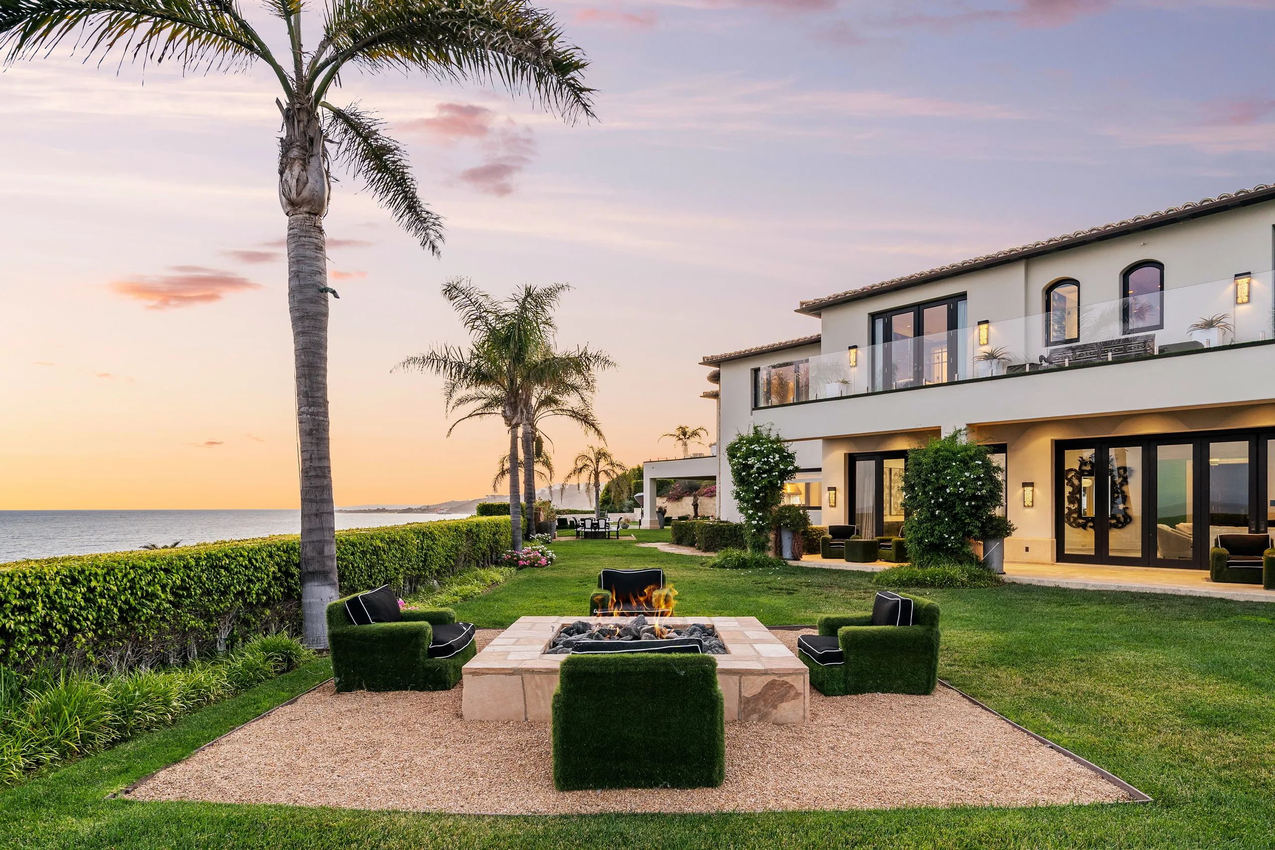 Lit fire pit ringed by turf armchairs on the ocean-front lawn, villa windows glowing at sunset