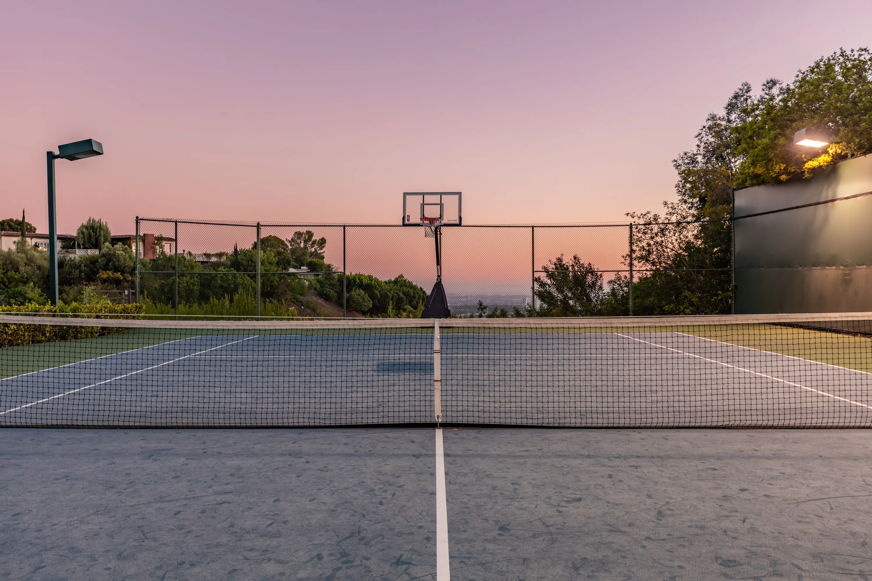 Full-size private tennis court with basketball hoop and city lights beyond the fence at dusk