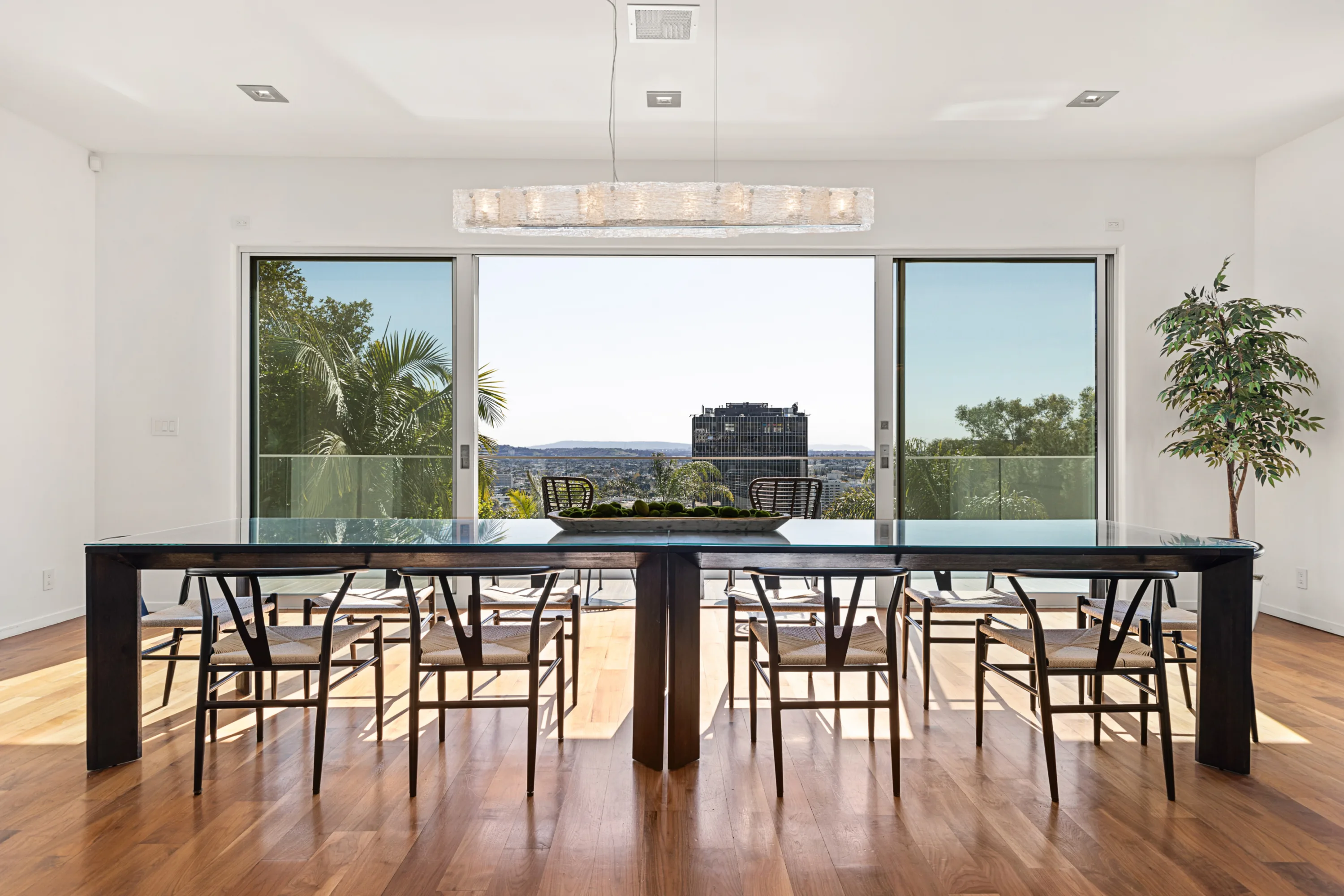 Long glass-topped dining table for twelve framed by a glowing linear chandelier and panoramic city outlook