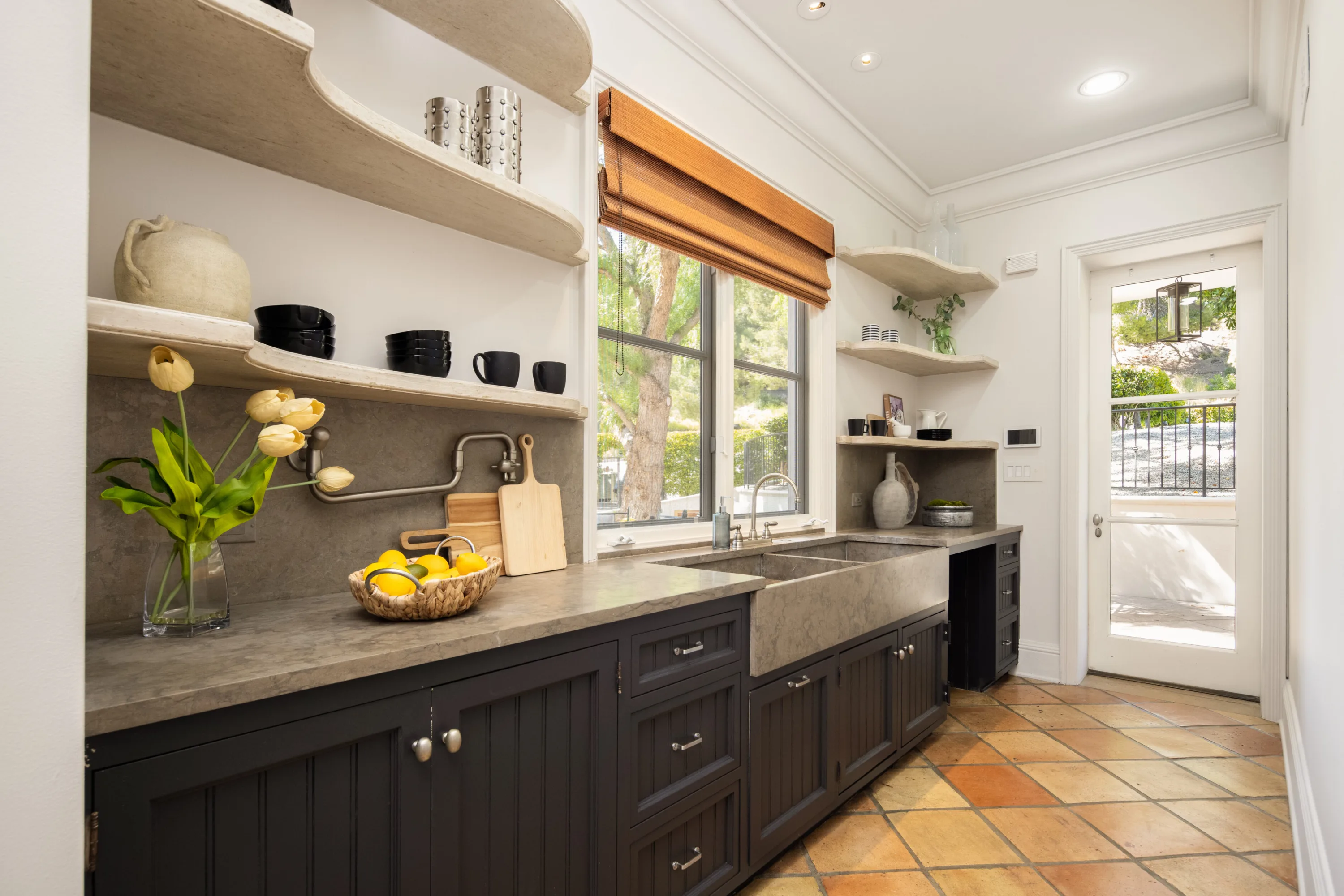 Butler's pantry with dark cabinetry, stone farmhouse sink, open shelving and terracotta tile floor
