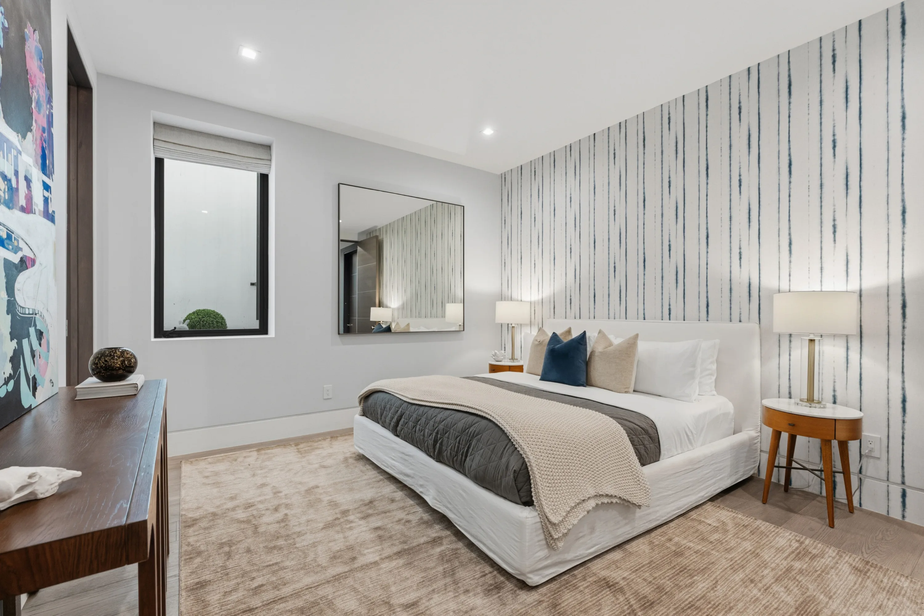 Bright bedroom with striped feature wall, king bed dressed in layered neutrals and colourful canvas by the door