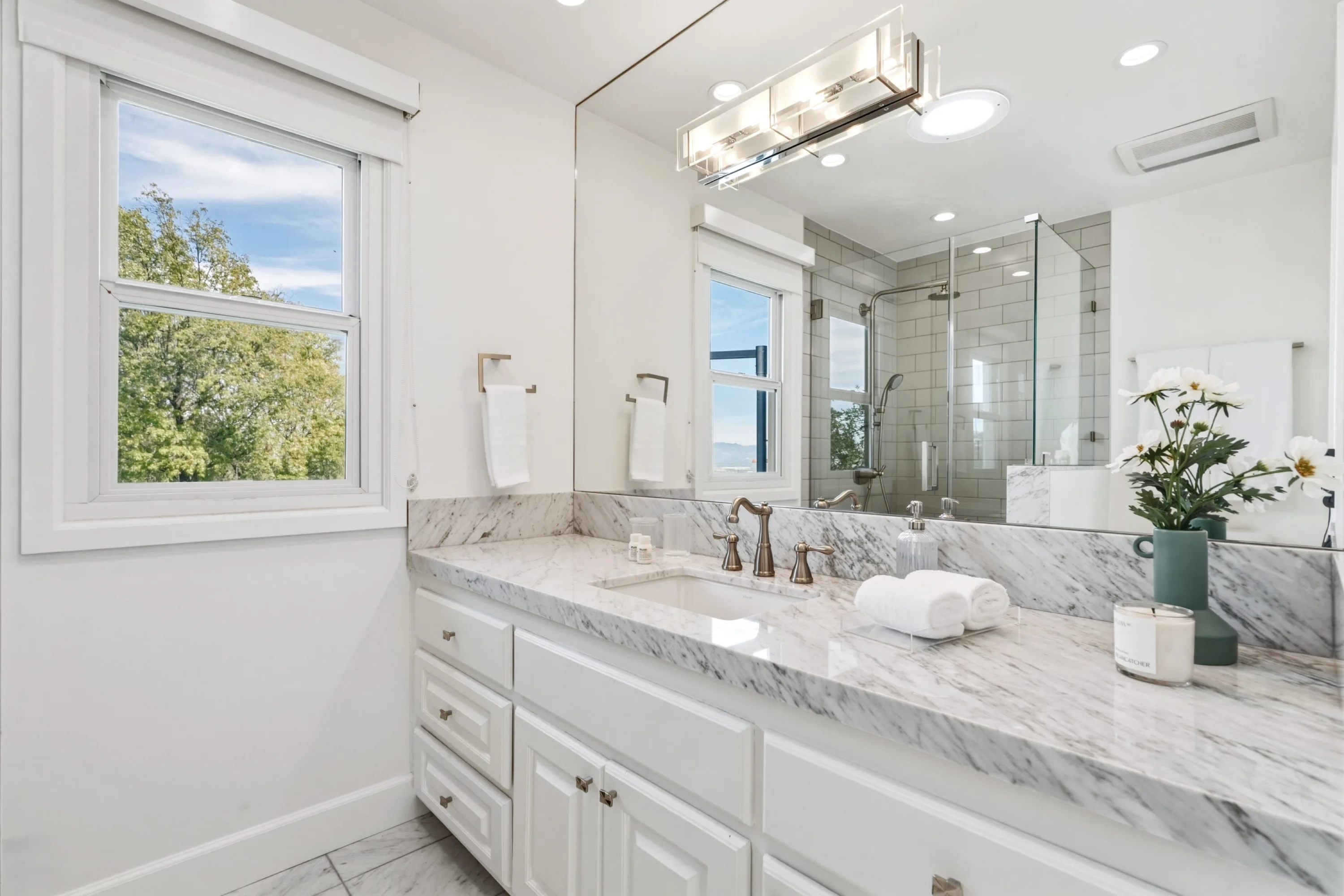 Double-window bathroom vanity in marble, with treetop outlook and glass shower reflected in the mirror