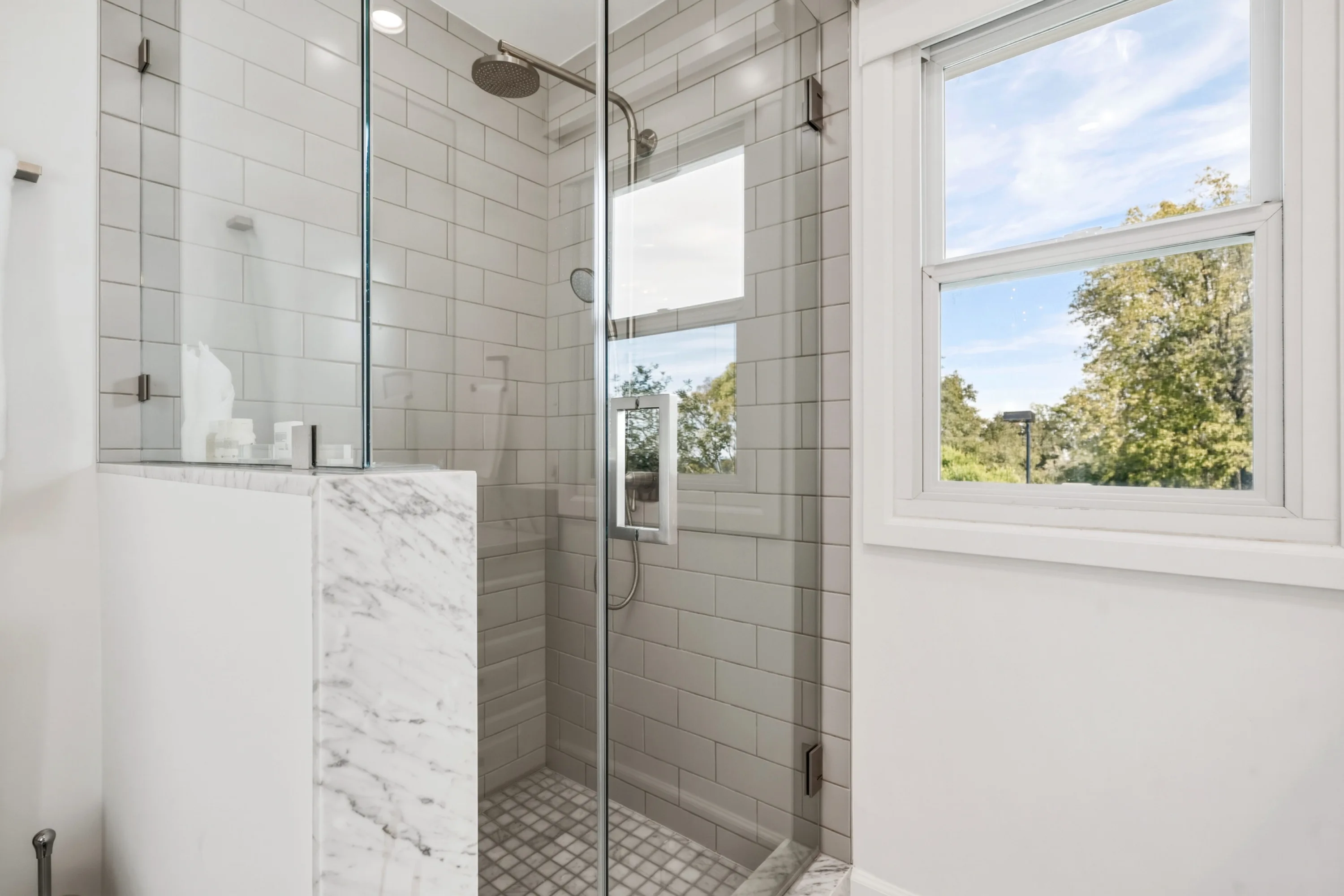 Glass shower with marble half-wall and window framing treetops and blue sky