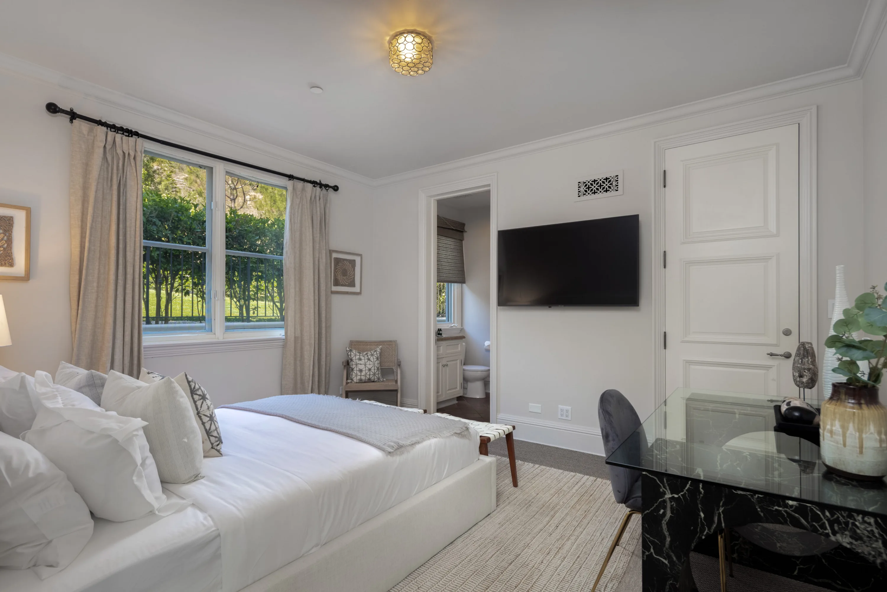 Guest bedroom with white linen bed, glass desk, wall-mounted TV and garden view through the window