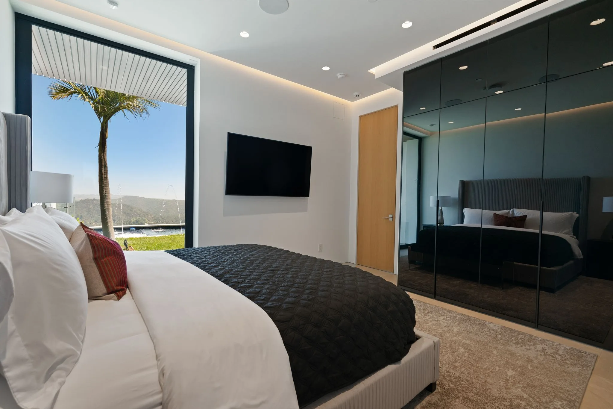 Bedroom opening to a lawn and canyon ridgeline through a full-height window, mirrored wardrobe wall opposite