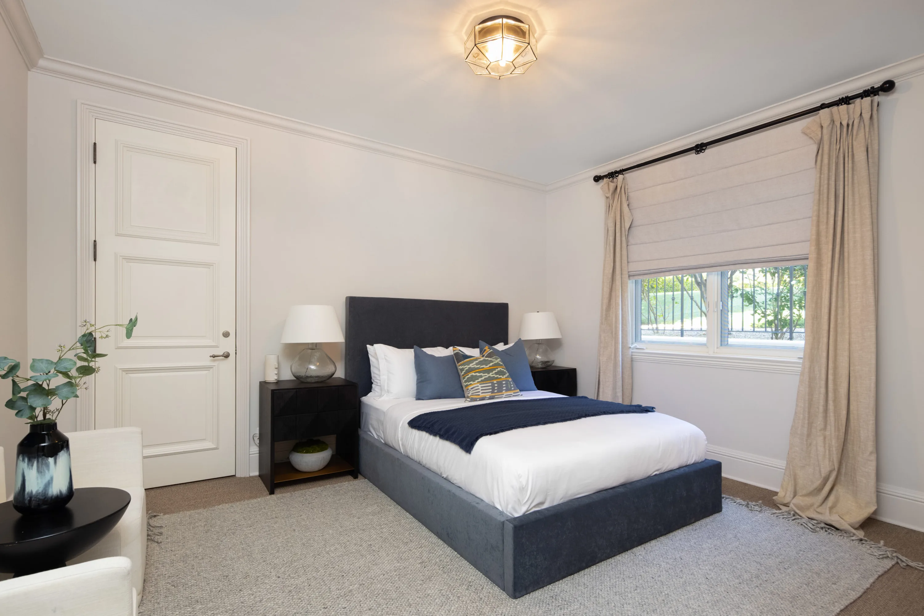 Navy upholstered bed and matching lamps in a calm guest room with curtained window onto the lawn