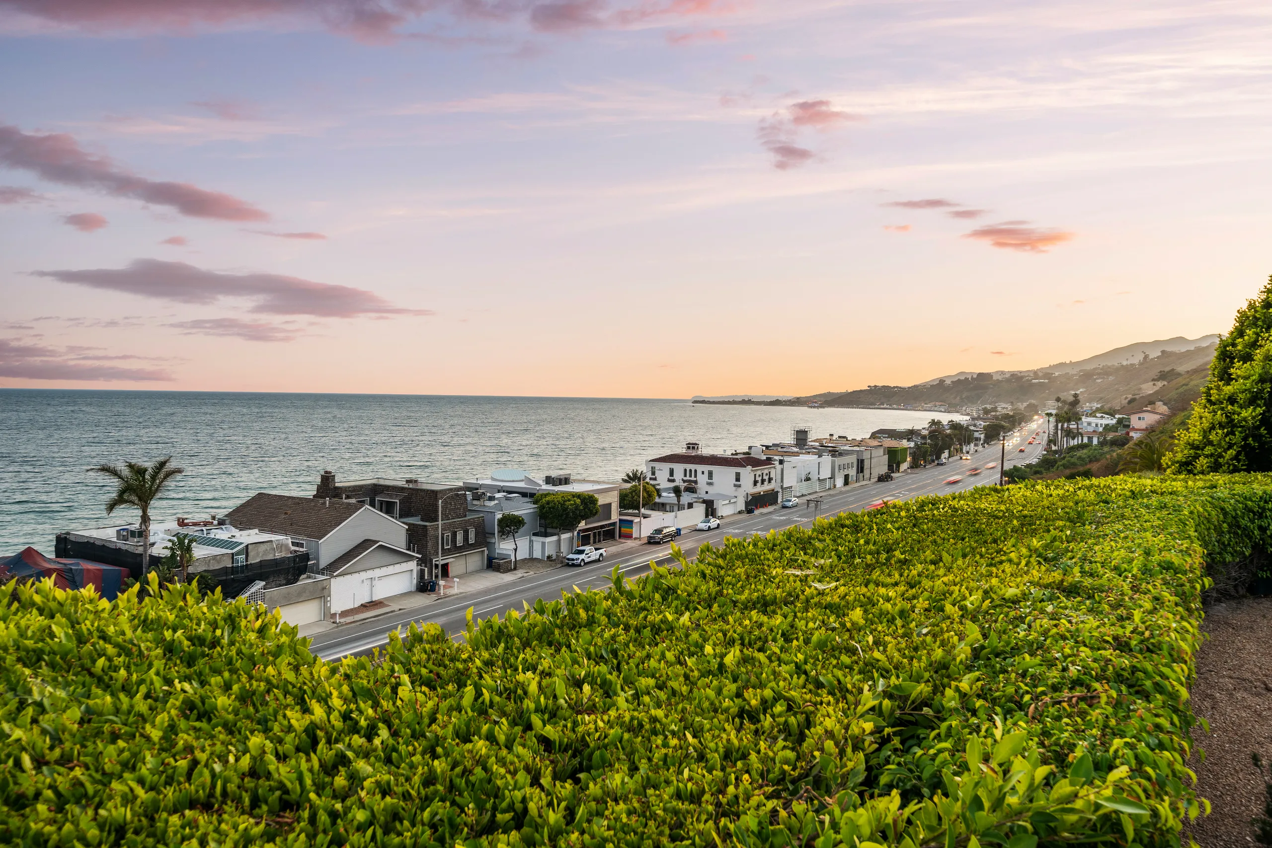 Sunset view over the hedge to the shoreline houses, coast road and pink-lit Pacific